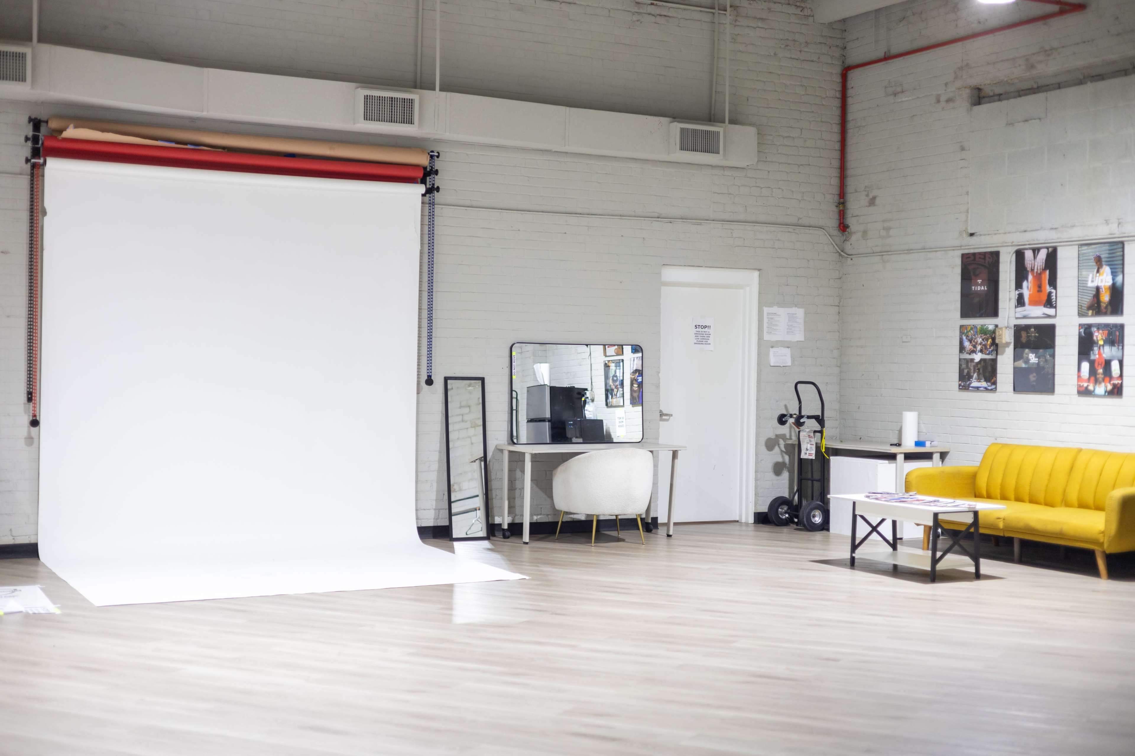 The image shows a spacious photography studio featuring a white backdrop, a mirror, a table, a yellow sofa, and posters on the walls.
