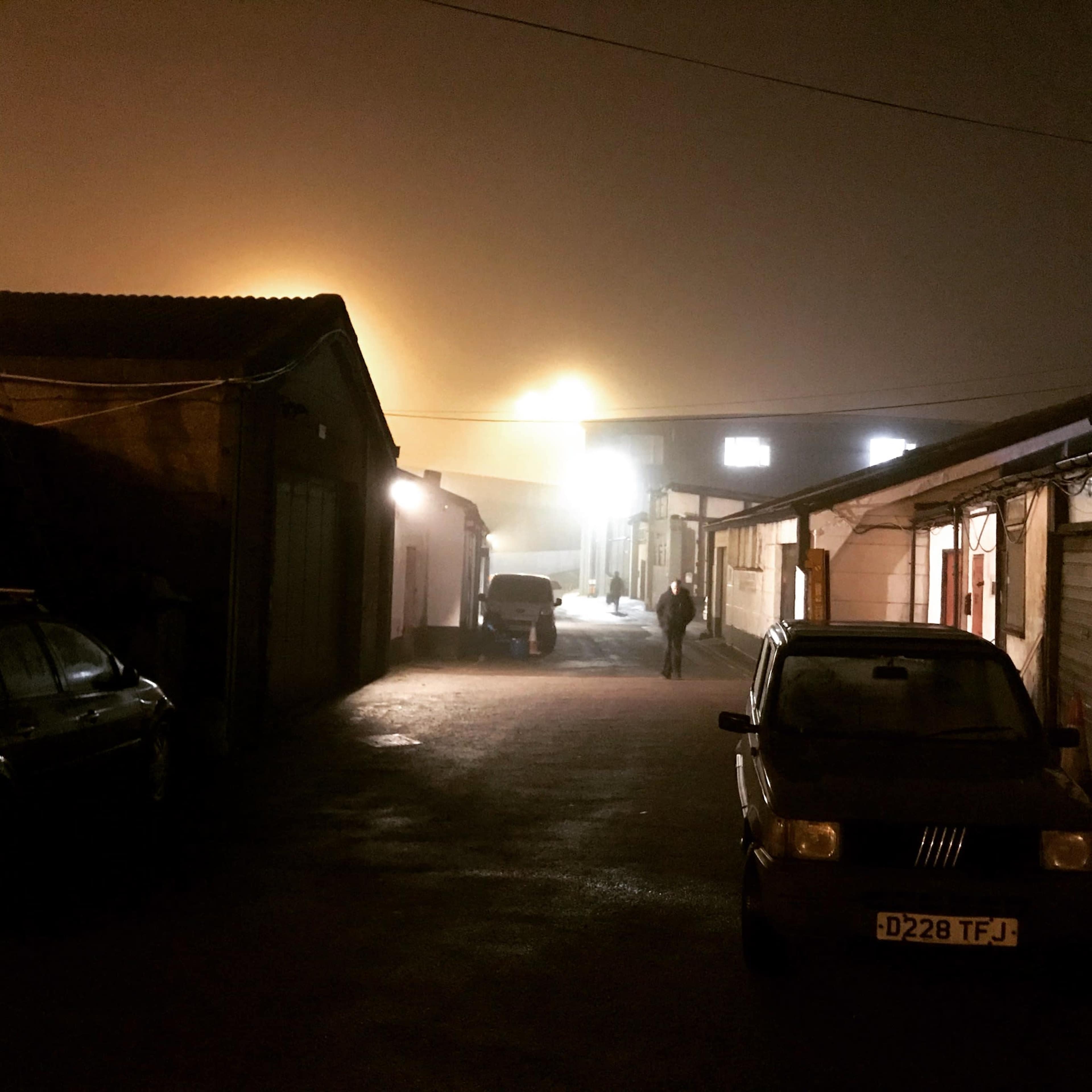 A person walks down a dimly lit alley surrounded by industrial buildings on a foggy night.