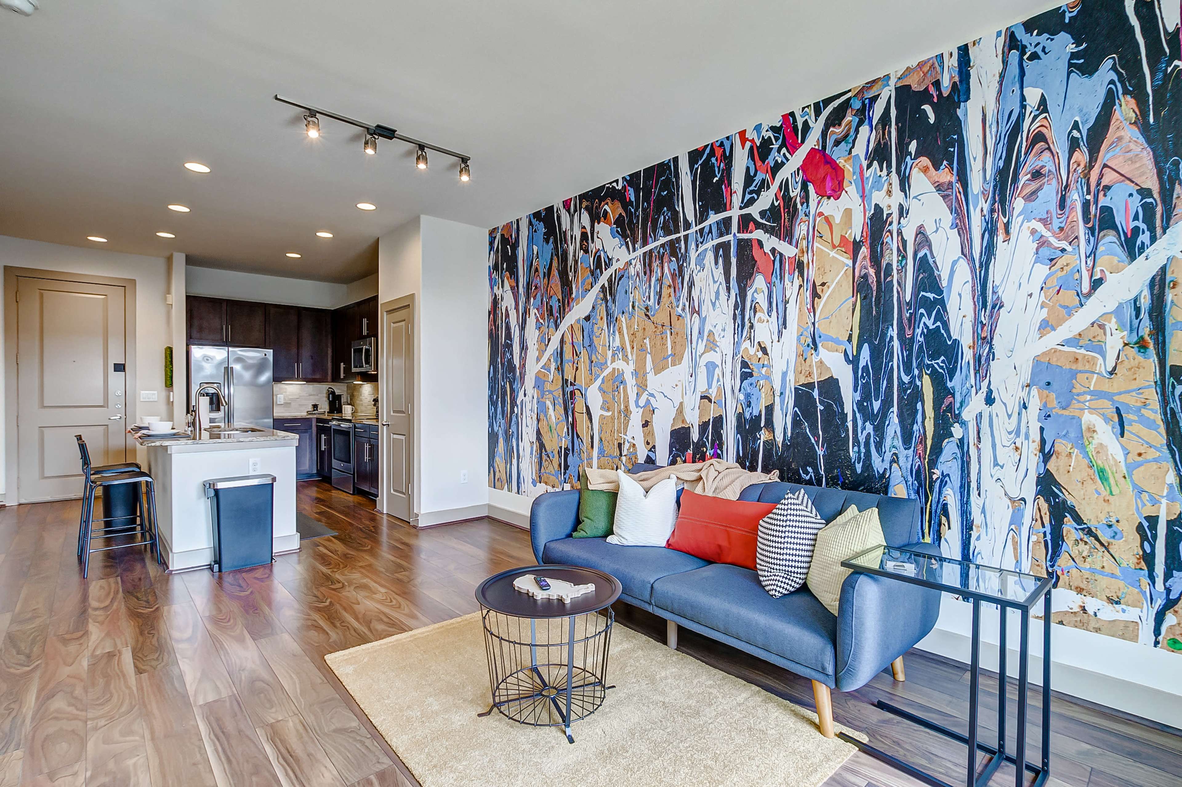 A modern living room with a blue sofa, a round coffee table, and a large, colorful mural on one wall, alongside a kitchen area in the background.
