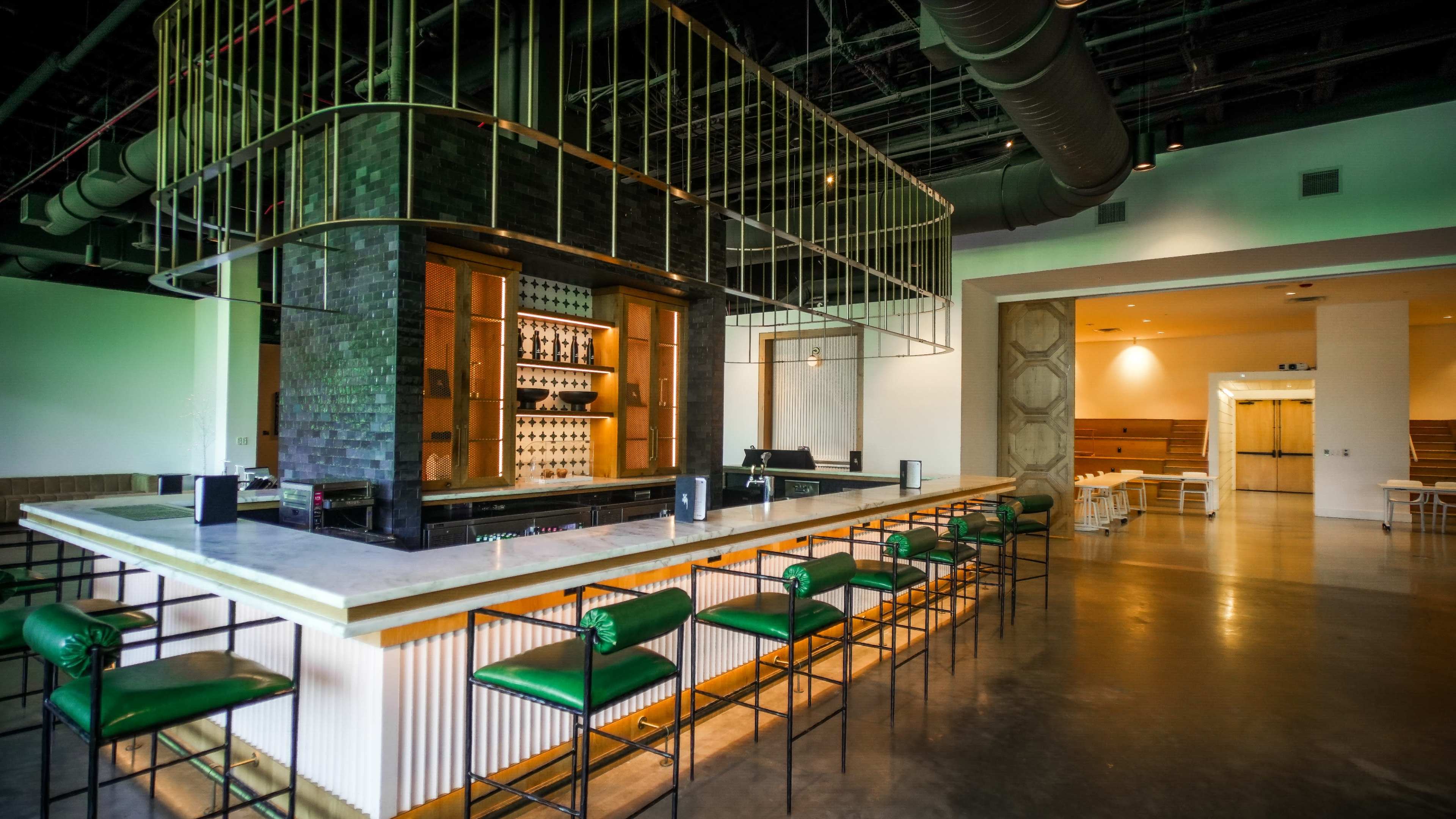 The image shows a modern bar area with a white marble countertop, green leather bar stools, and a backdrop featuring shelves lined with bottles.