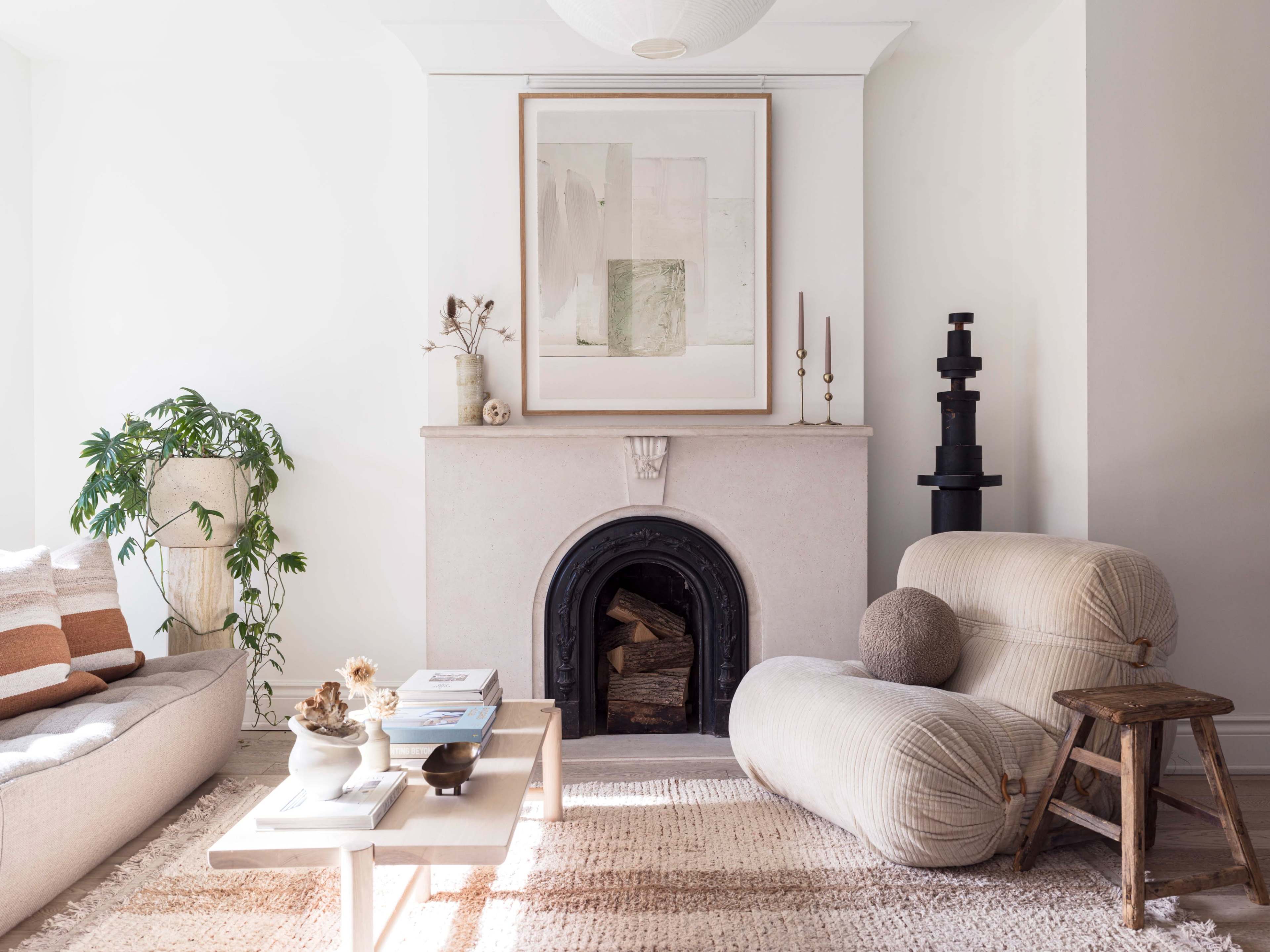 A bright living room features a fireplace with a framed artwork above it, a sofa with decorative pillows, a coffee table with books, and a wooden stool beside a soft, sculptural chair.