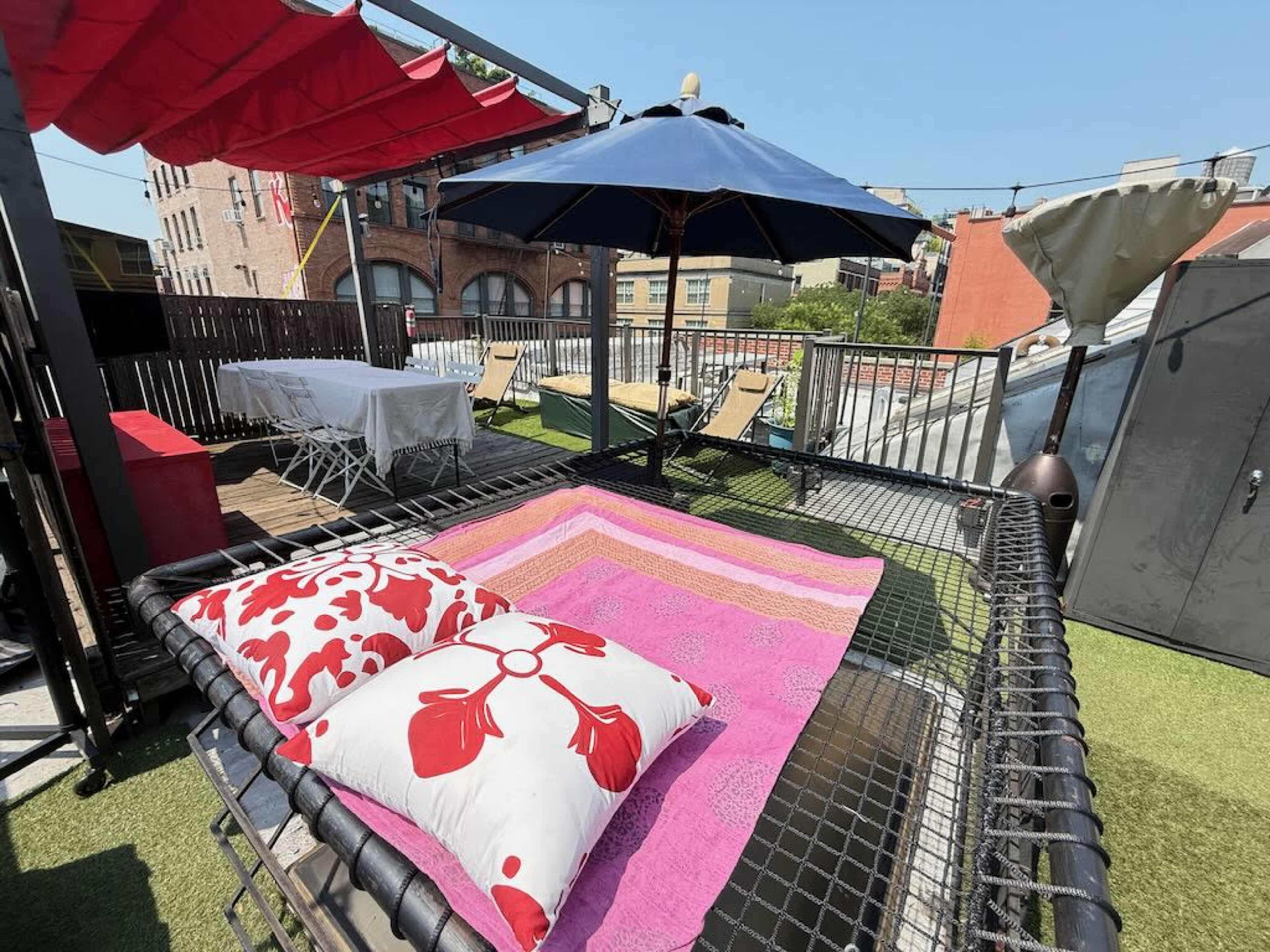 The image shows a rooftop area featuring a large hammock with decorative pillows, lounge chairs, and an umbrella under a clear blue sky.