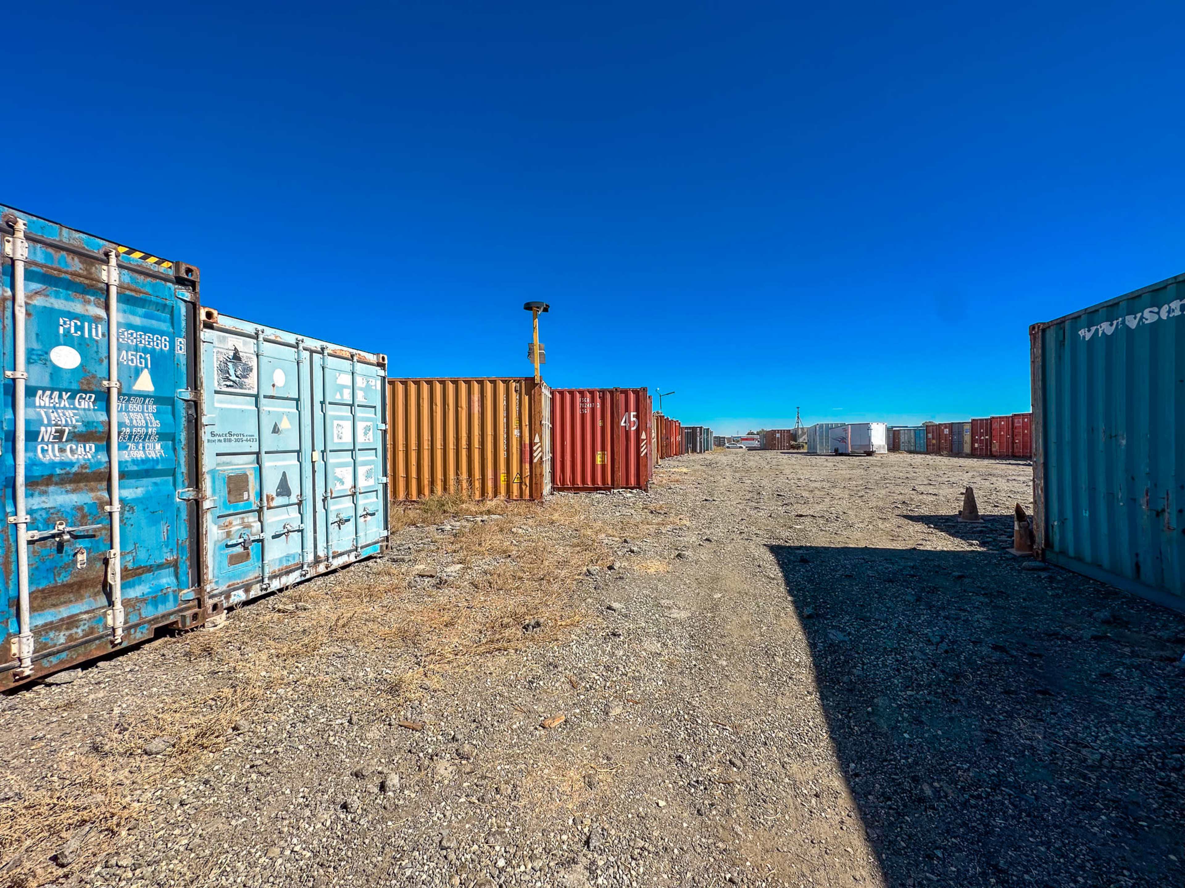The image shows a row of shipping containers in various colors lined along a gravel path under a clear blue sky.