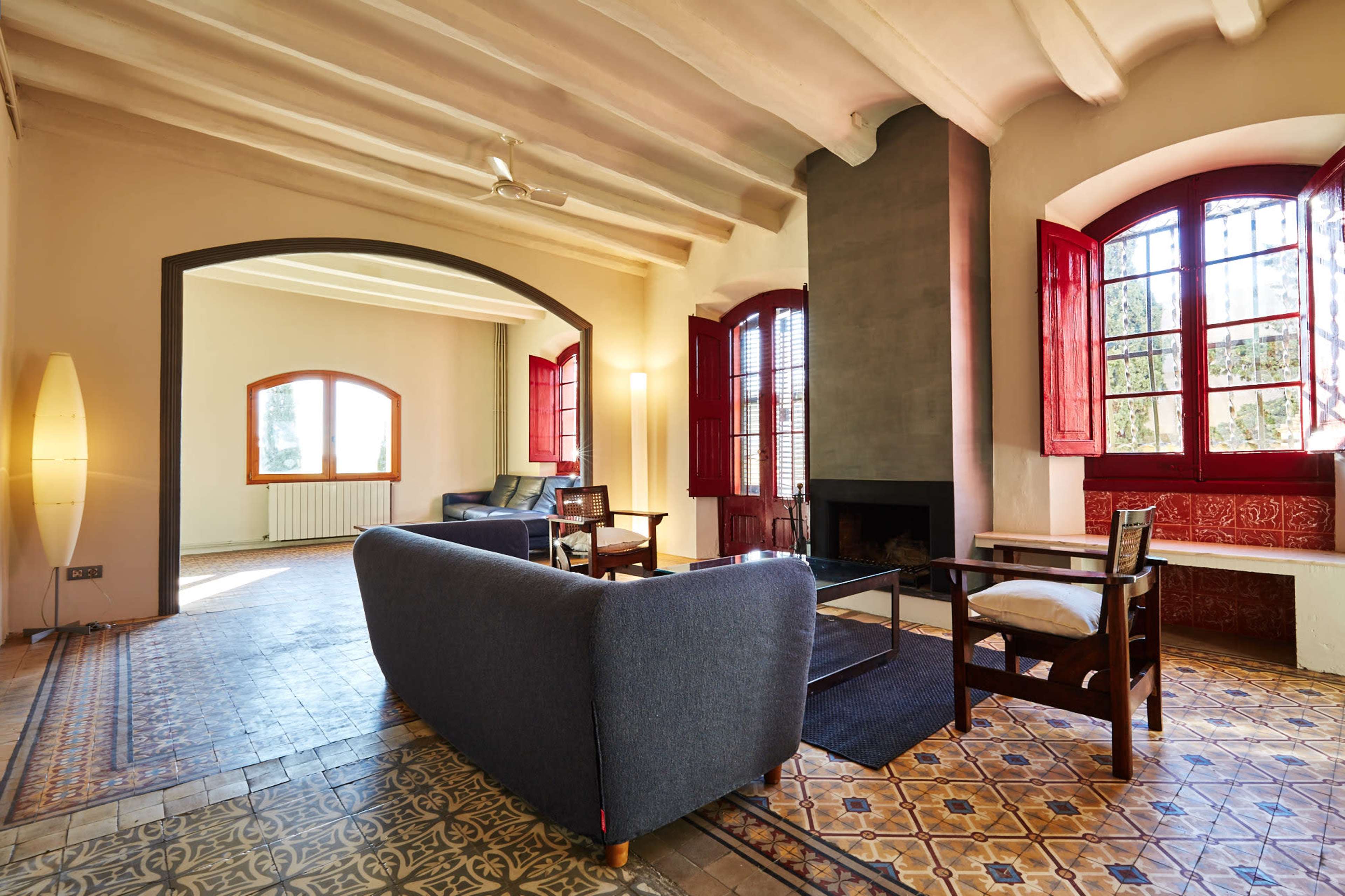 A spacious living area featuring a gray sofa, wooden furniture, a fireplace, and large windows with red shutters allowing natural light to enter.