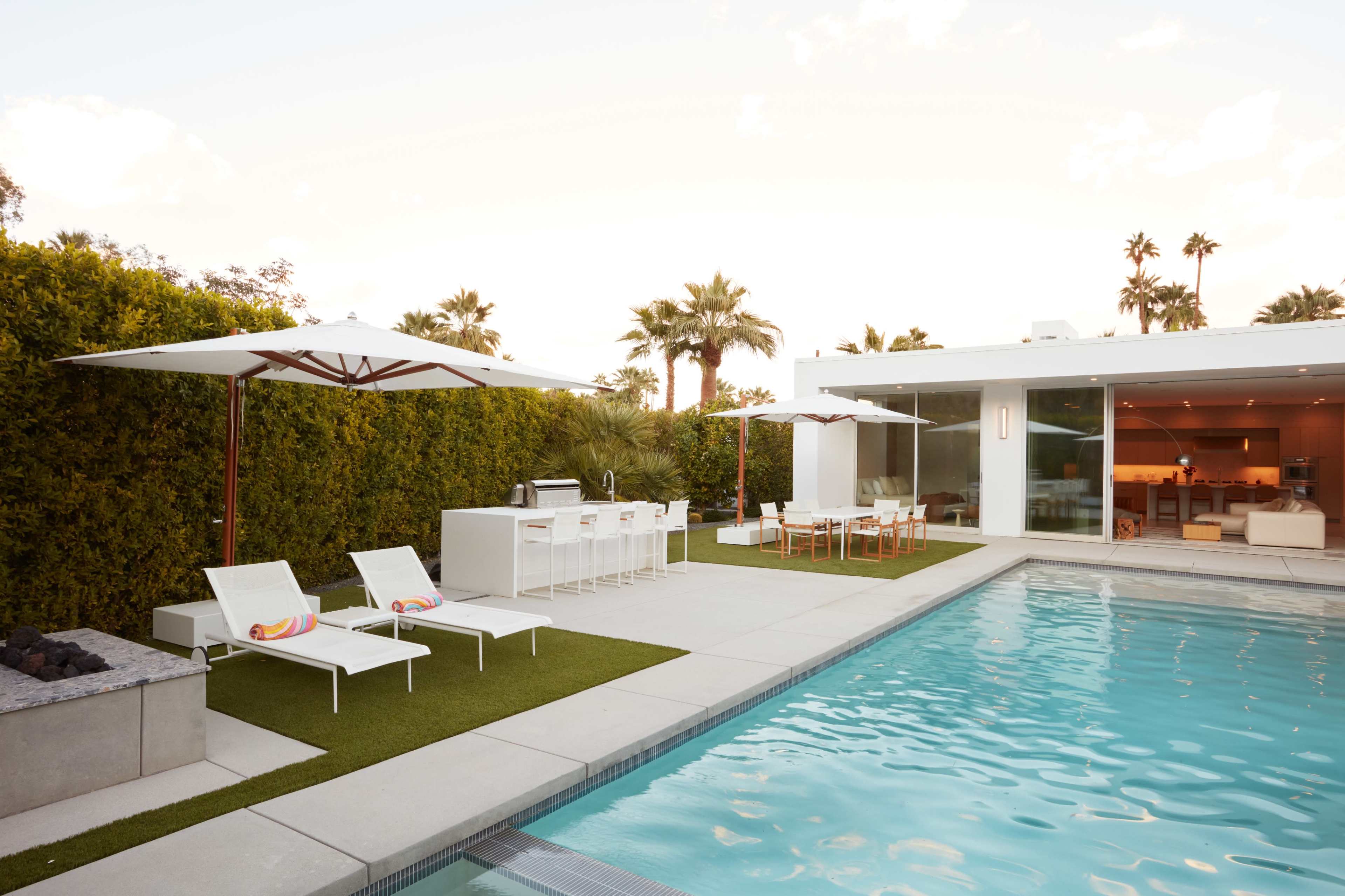 The image depicts a modern backyard with a swimming pool, lounge chairs, a barbecue area, and an outdoor dining space surrounded by palm trees.