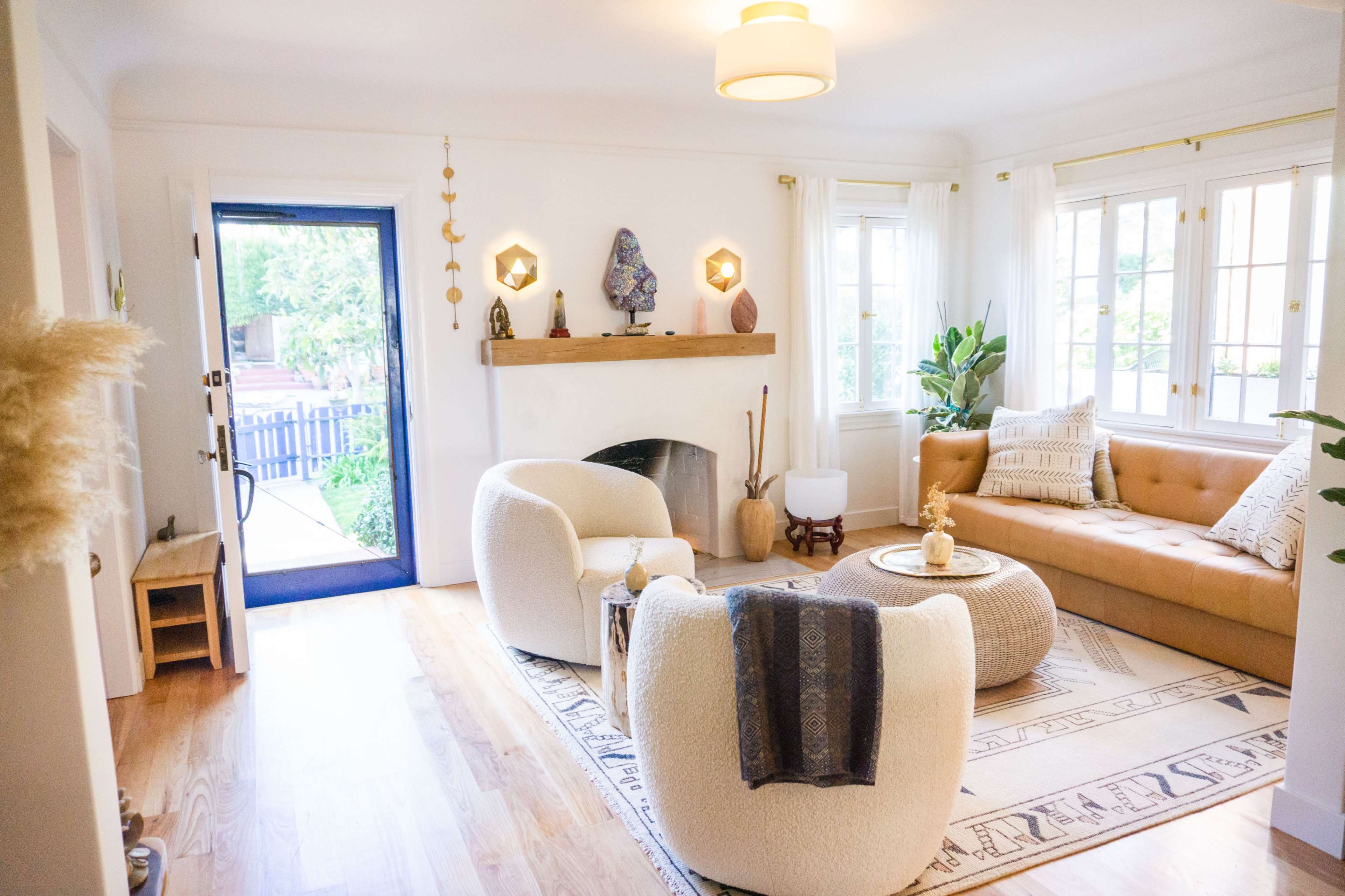 A cozy living room features a tan sofa, two white armchairs, a round coffee table, and large windows overlooking a blue door and outdoor area.