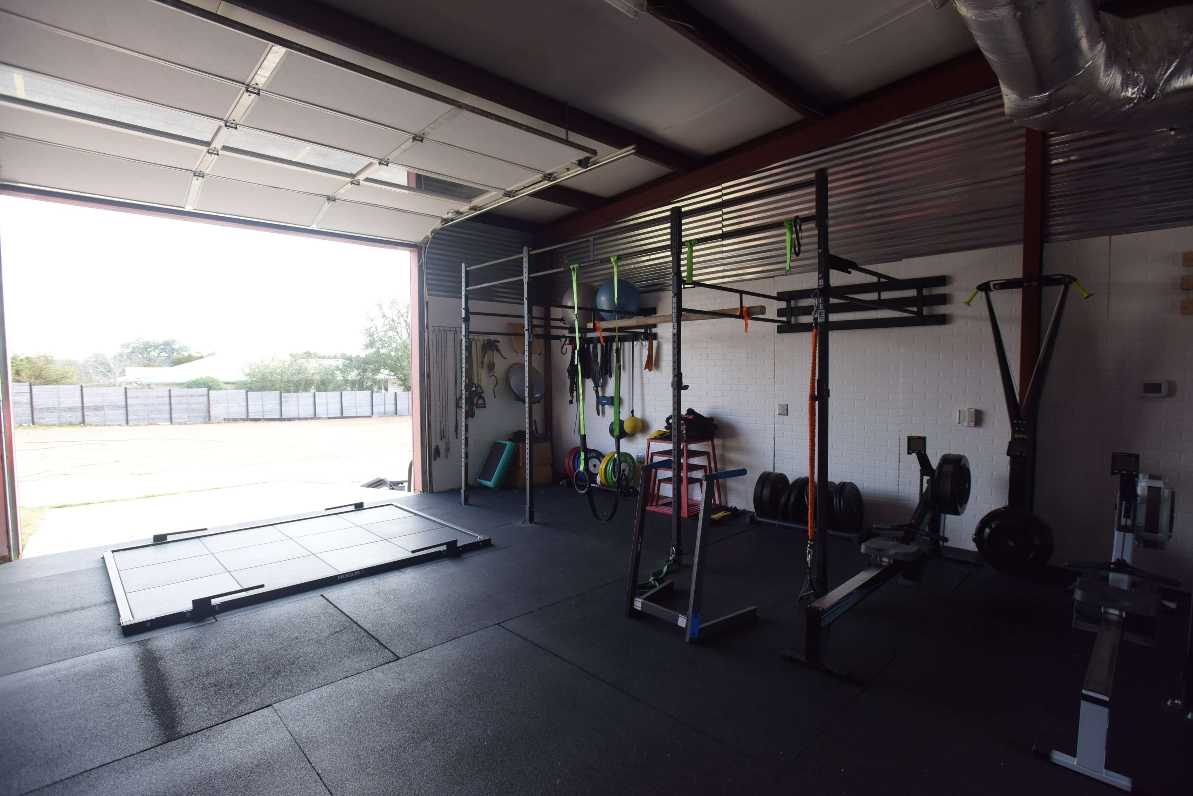 A spacious gym with various exercise equipment, including a squat rack, weights, and rowing machines, set up inside a garage with an open door allowing natural light in.