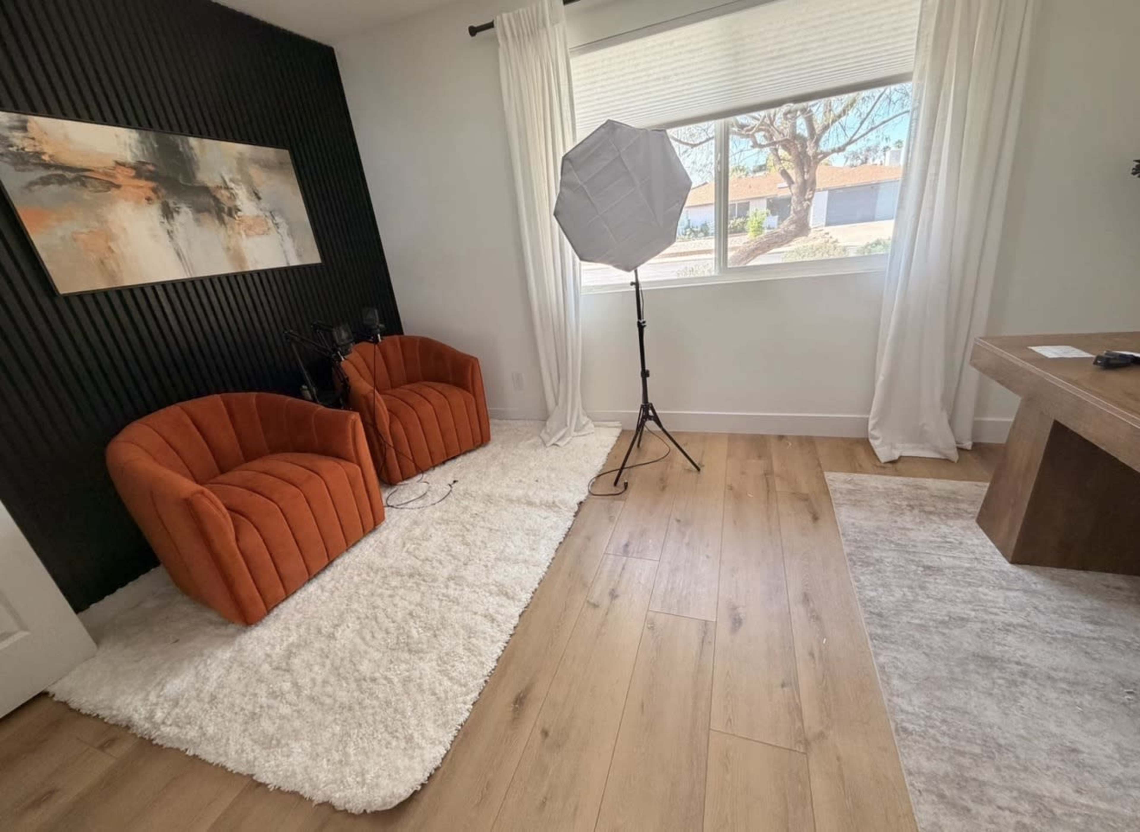 The image shows a modern room featuring two orange velvet chairs, a soft white area rug, a large window with sheer curtains, and a photography light setup.