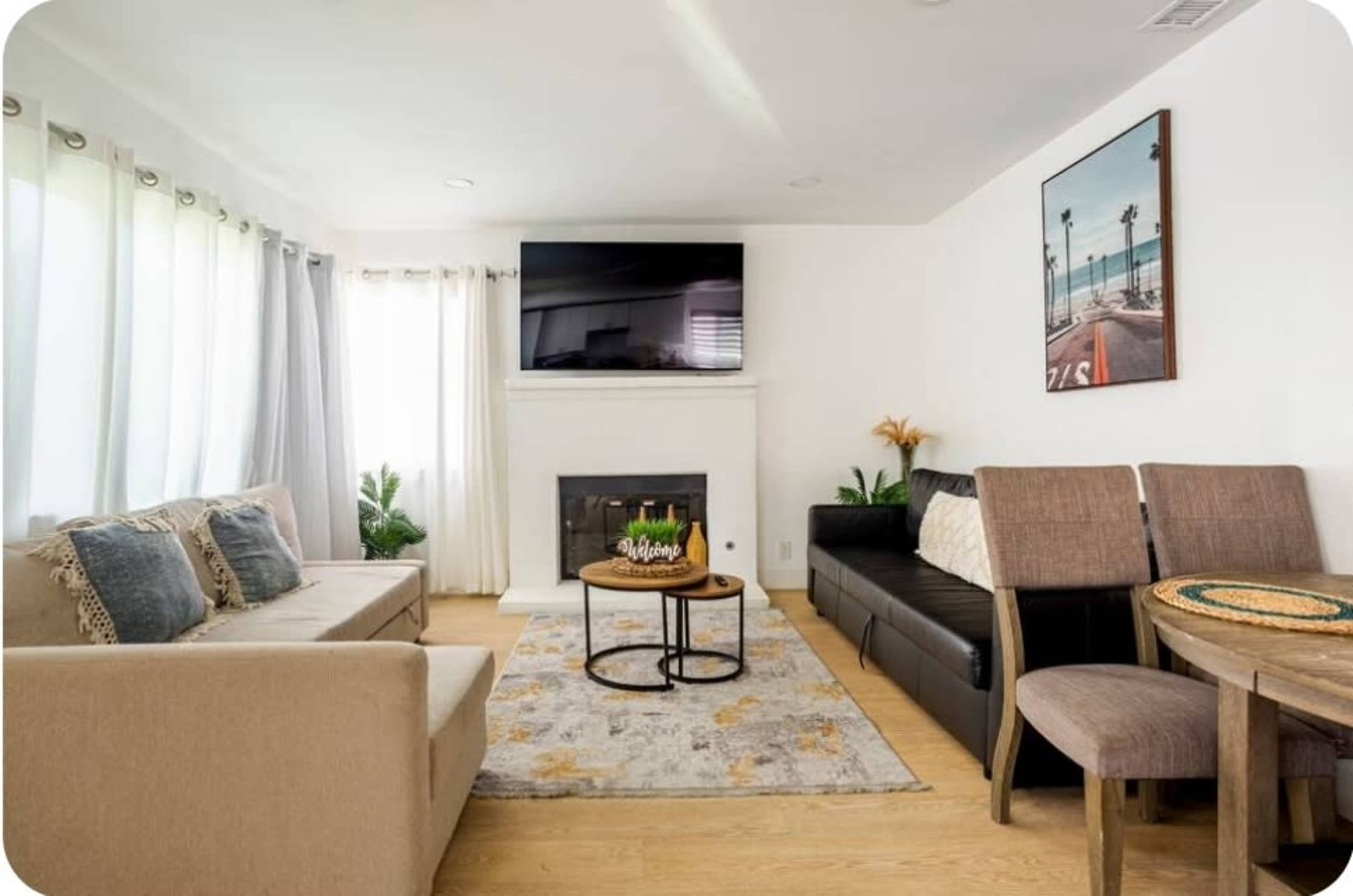 The living room features a light-colored sofa and a dark couch arranged around a circular coffee table on a patterned rug, with a fireplace and a television on the wall.