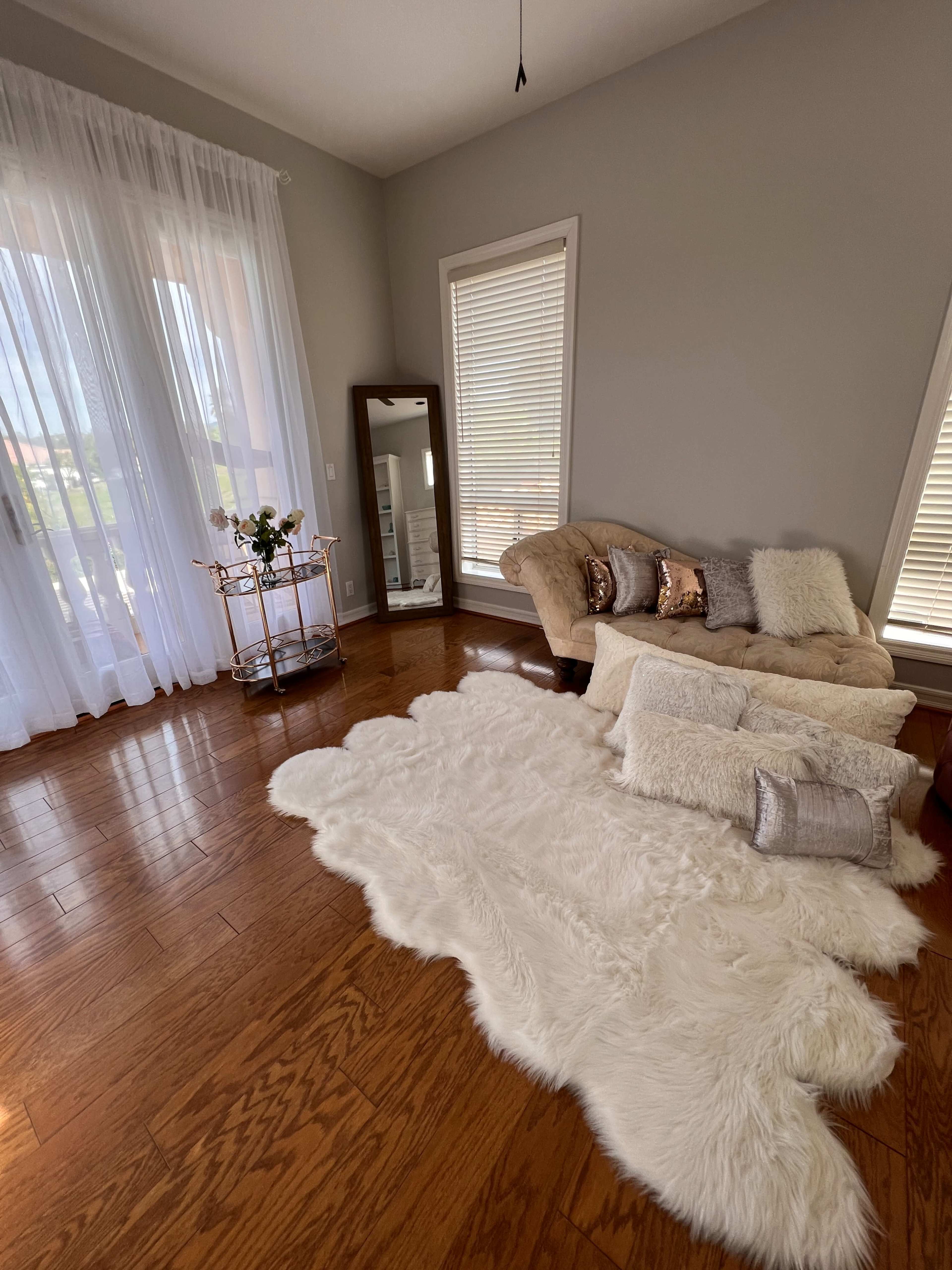 The image shows a well-lit living room with wooden flooring, featuring a beige couch, a large fluffy area rug, and a mirror beside a decorative cart.