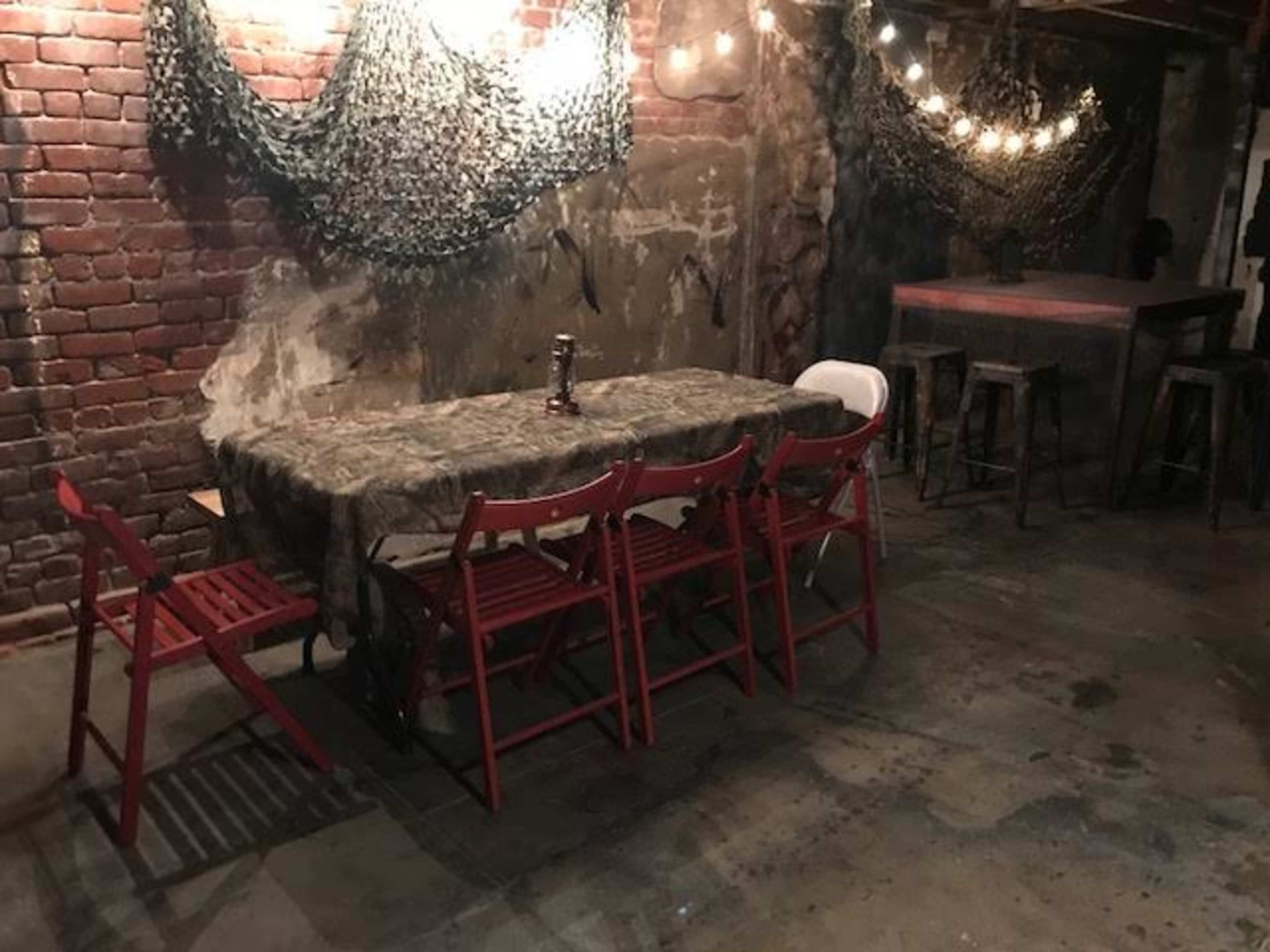 A rustic basement features a long table covered with a camouflage cloth, surrounded by red chairs, with dim lighting from hanging bulbs.