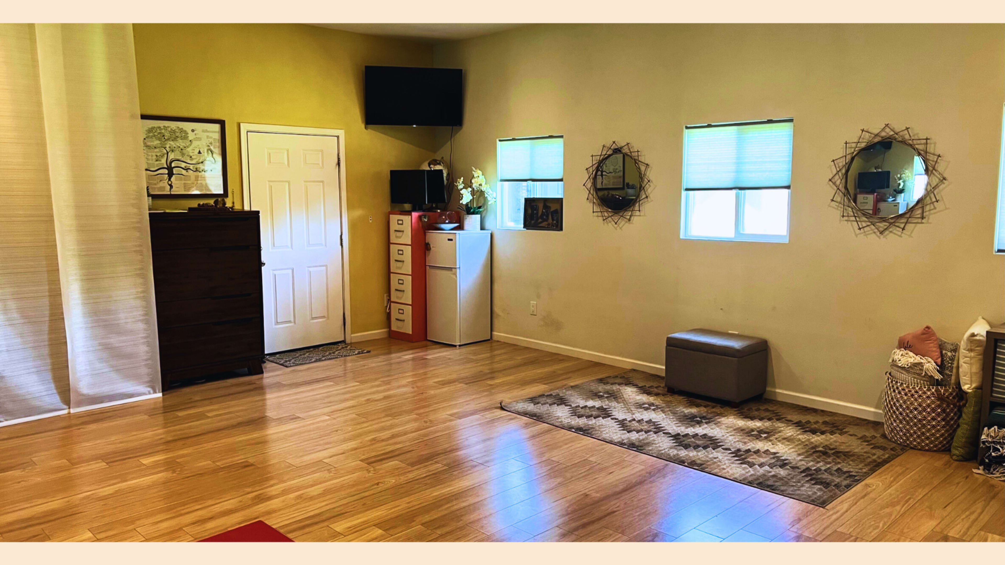 A spacious interior room with wooden flooring, featuring a door, two windows, a television mounted on the wall, and various furnishings including a bench and storage units.