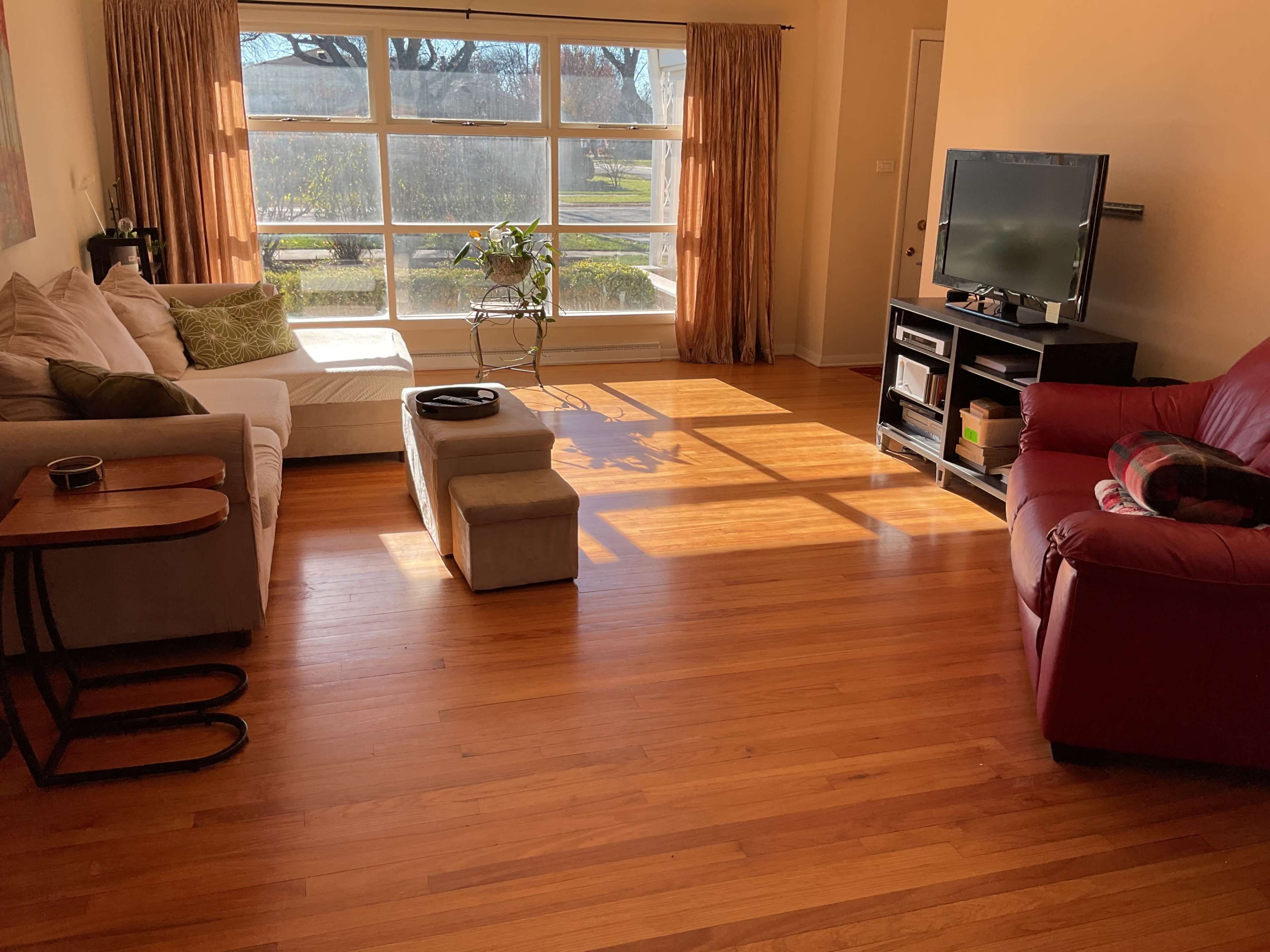 The image shows a living room with a large window allowing sunlight to illuminate the space, featuring a beige sofa, a red armchair, a small coffee table, and light-colored wooden flooring.