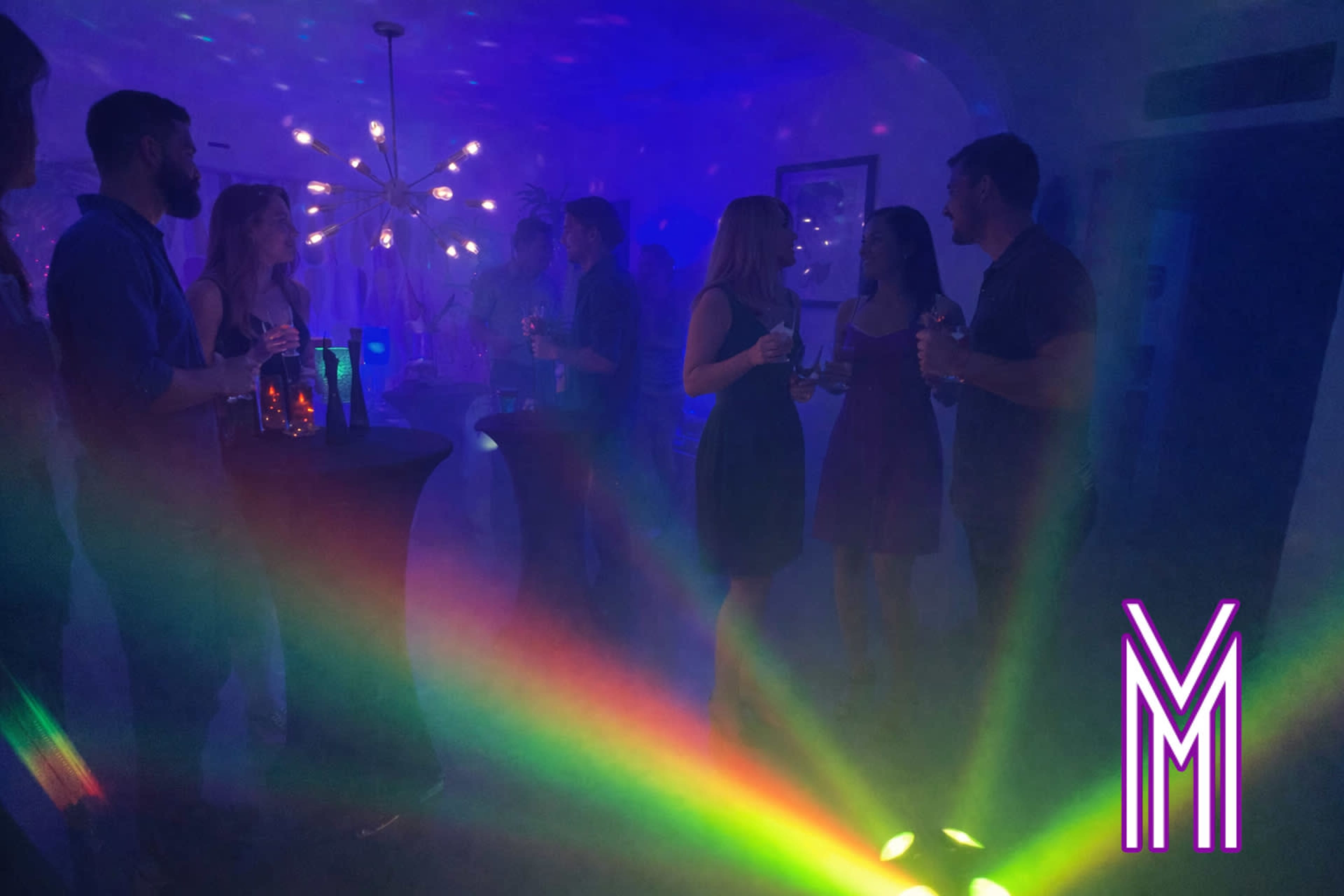 A group of people socializes at a vibrant party, illuminated by colorful lights and projections.