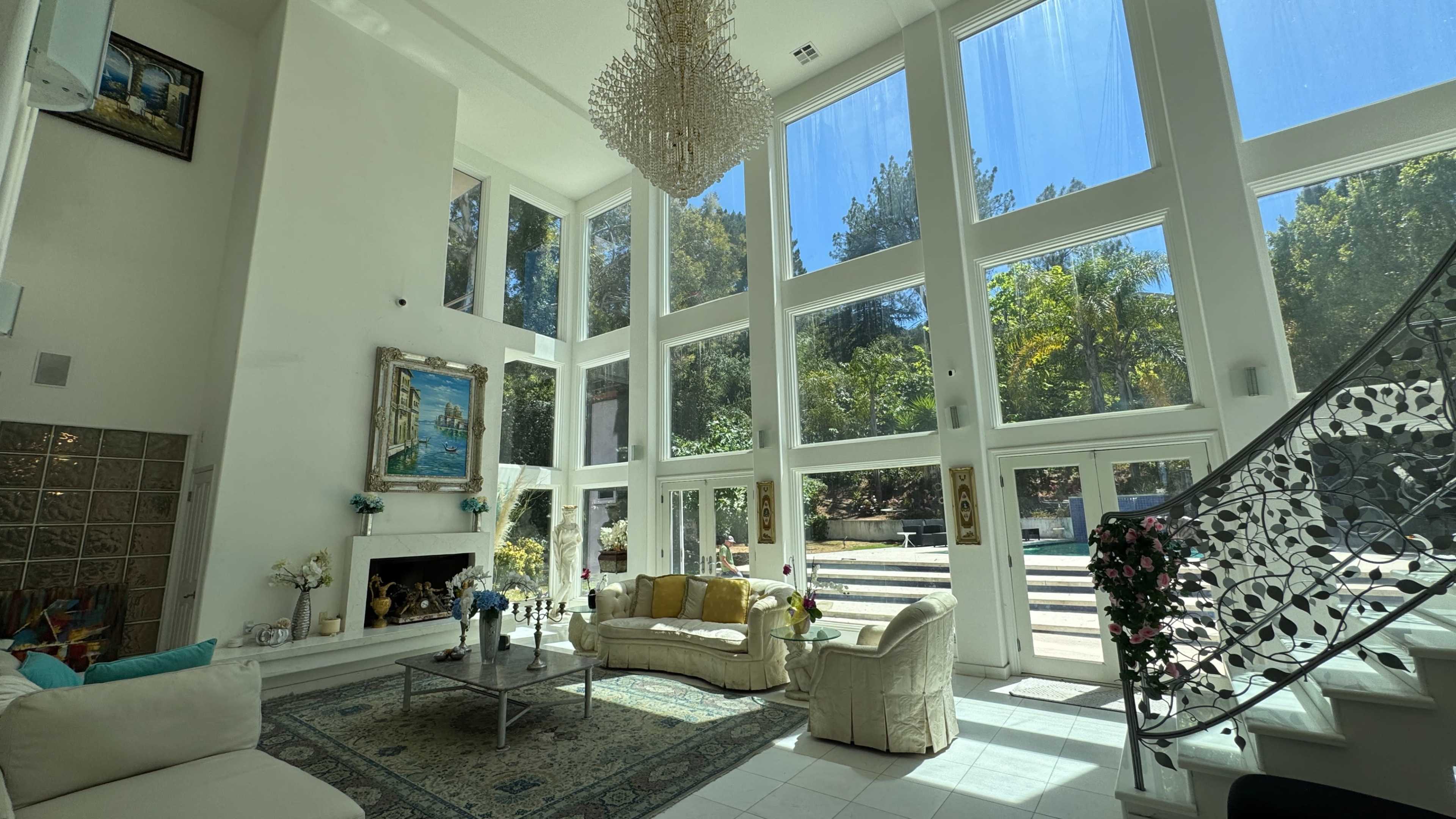 The image depicts a spacious living room with large windows, a chandelier, and a decorative staircase, featuring a white couch and a coffee table.