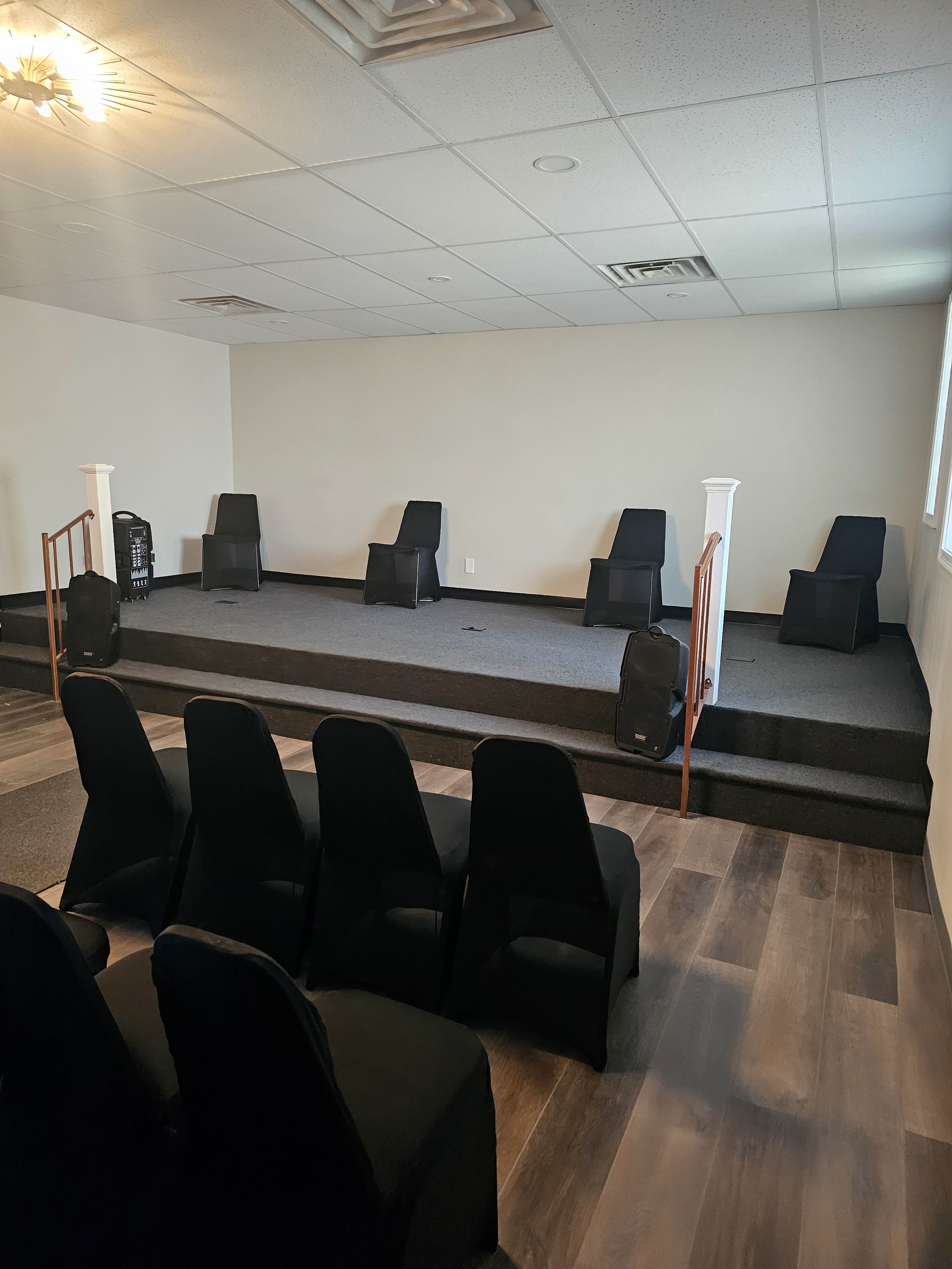 The image shows a small stage area with several black chairs arranged in rows and a few chairs positioned on the stage, all in a room with light-colored walls and a drop ceiling.
