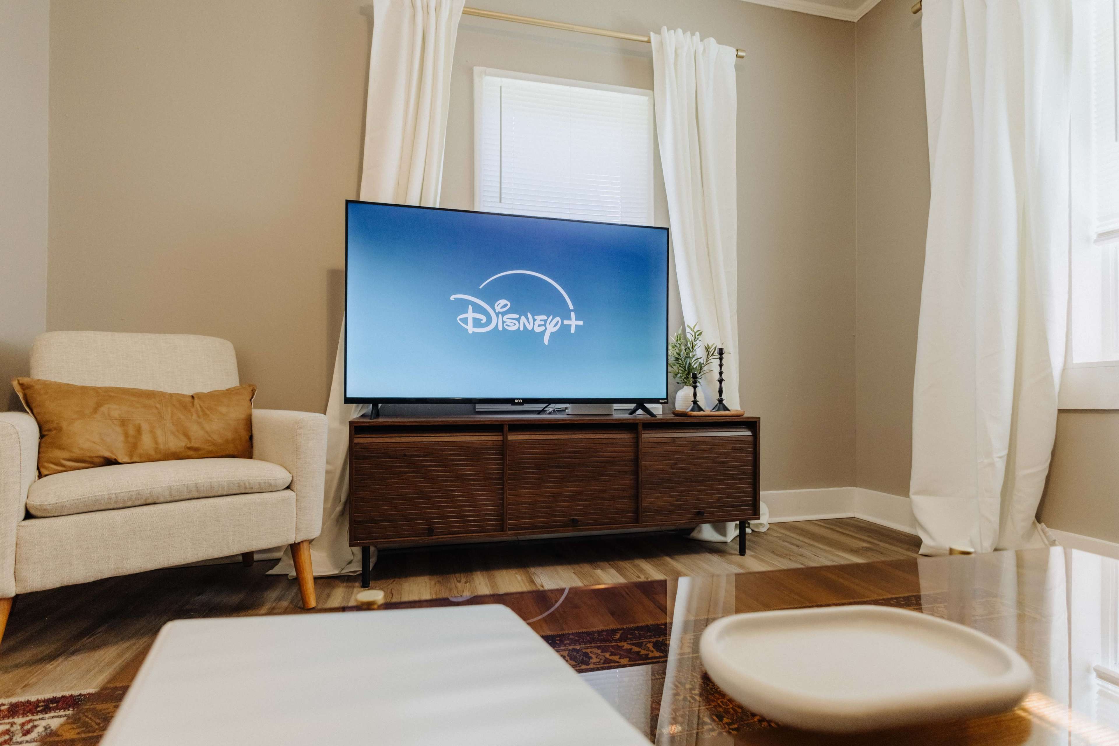 The image shows a living room with a television displaying the Disney+ logo, a beige armchair, and light-colored curtains.