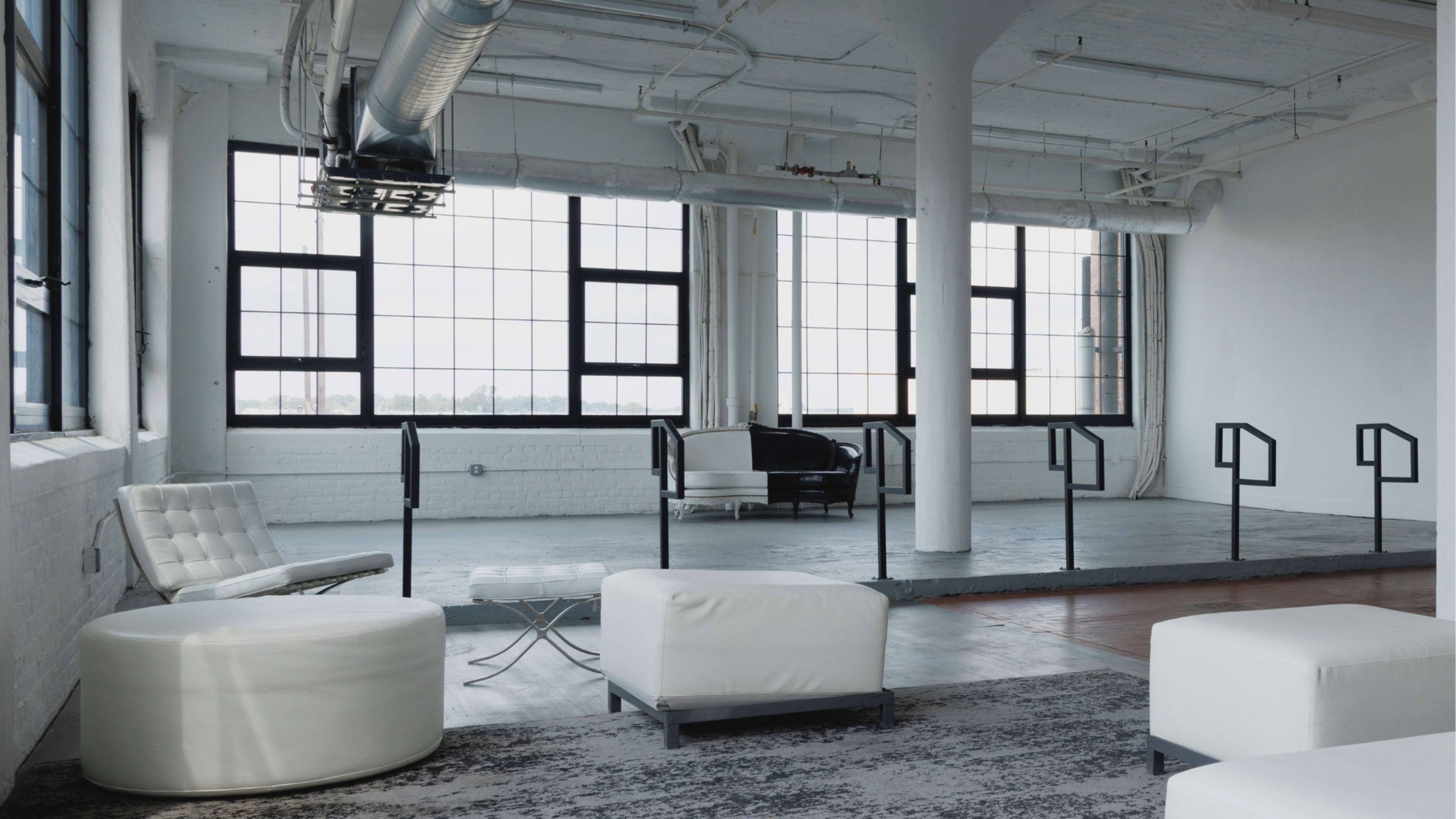 The image shows a spacious, modern interior with large windows, featuring minimalistic furniture arranged in a clean, open layout.