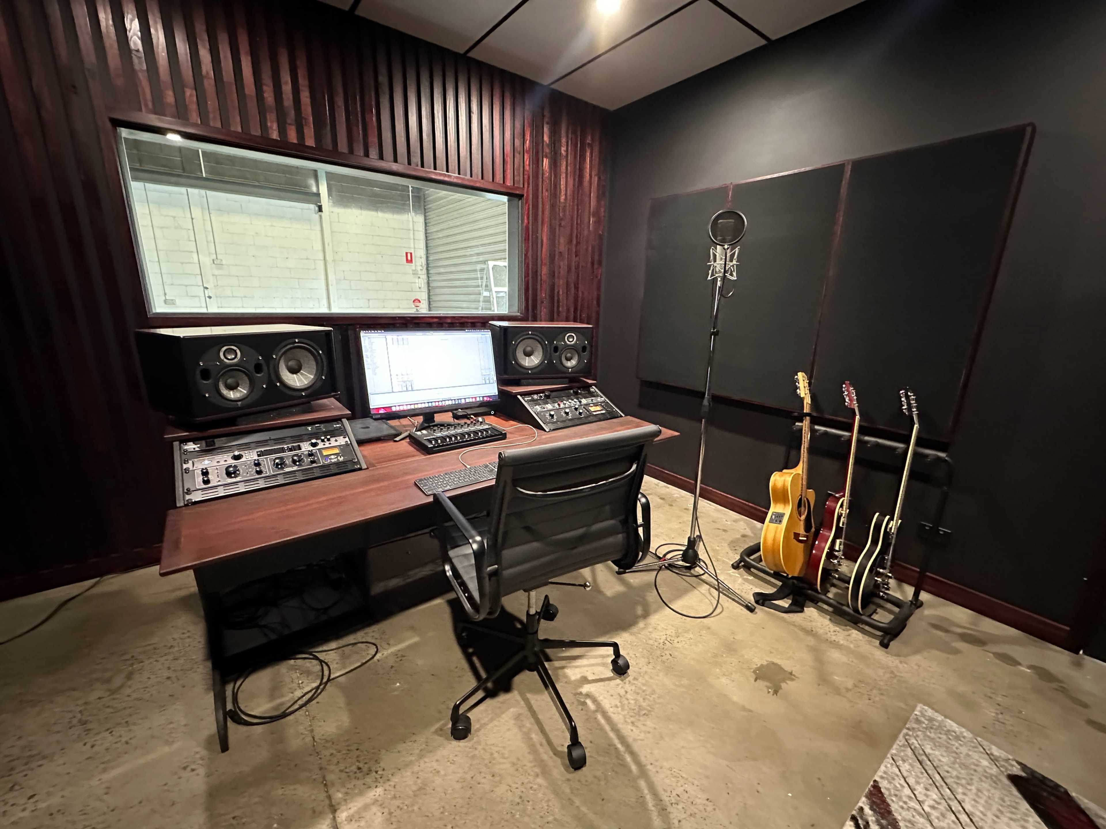 A music studio features a mixing desk with audio equipment, a computer screen, a microphone on a stand, and guitars stored in a rack.