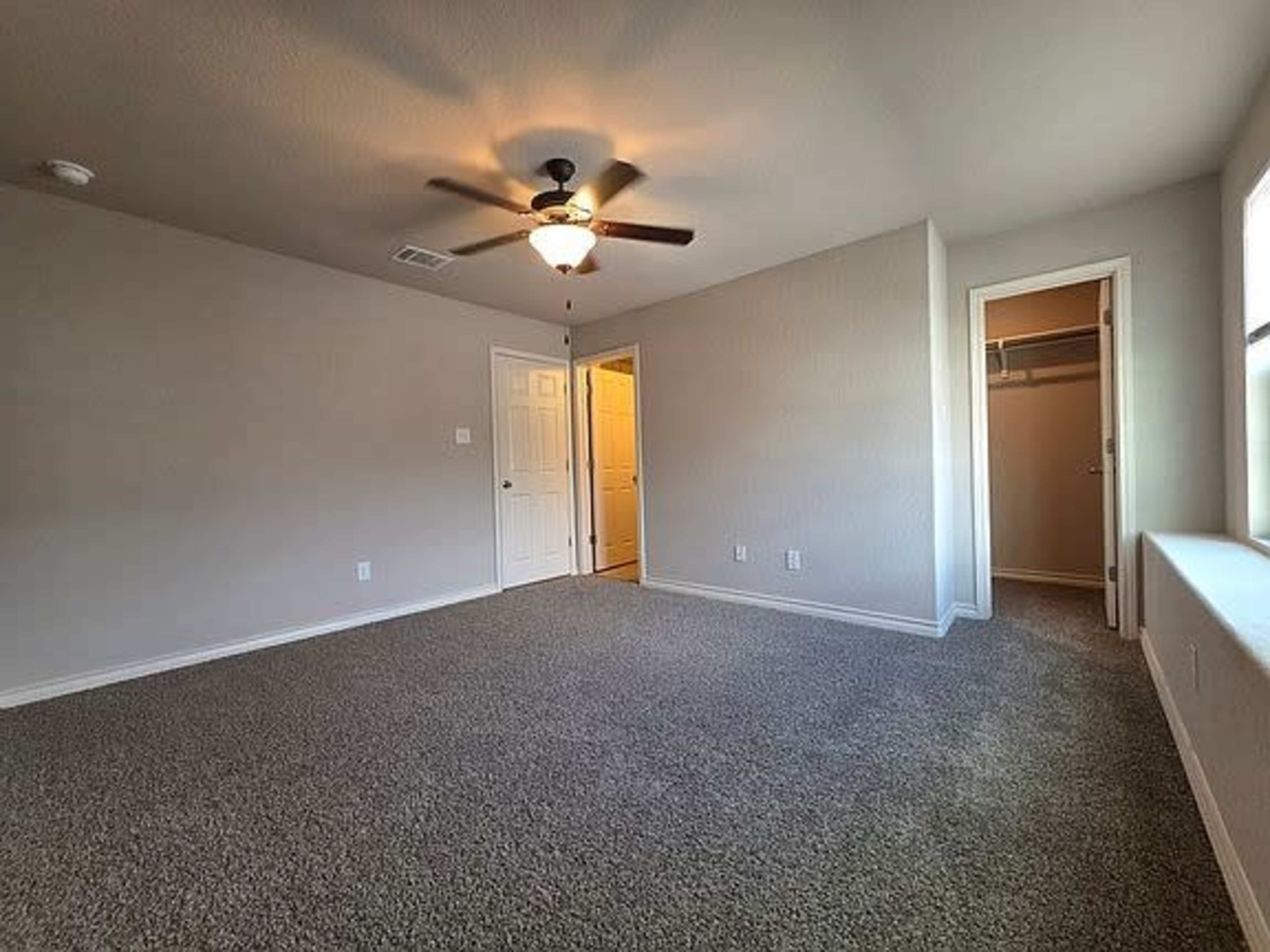The image shows a spacious, carpeted room with a ceiling fan and two doorways leading to adjacent spaces.