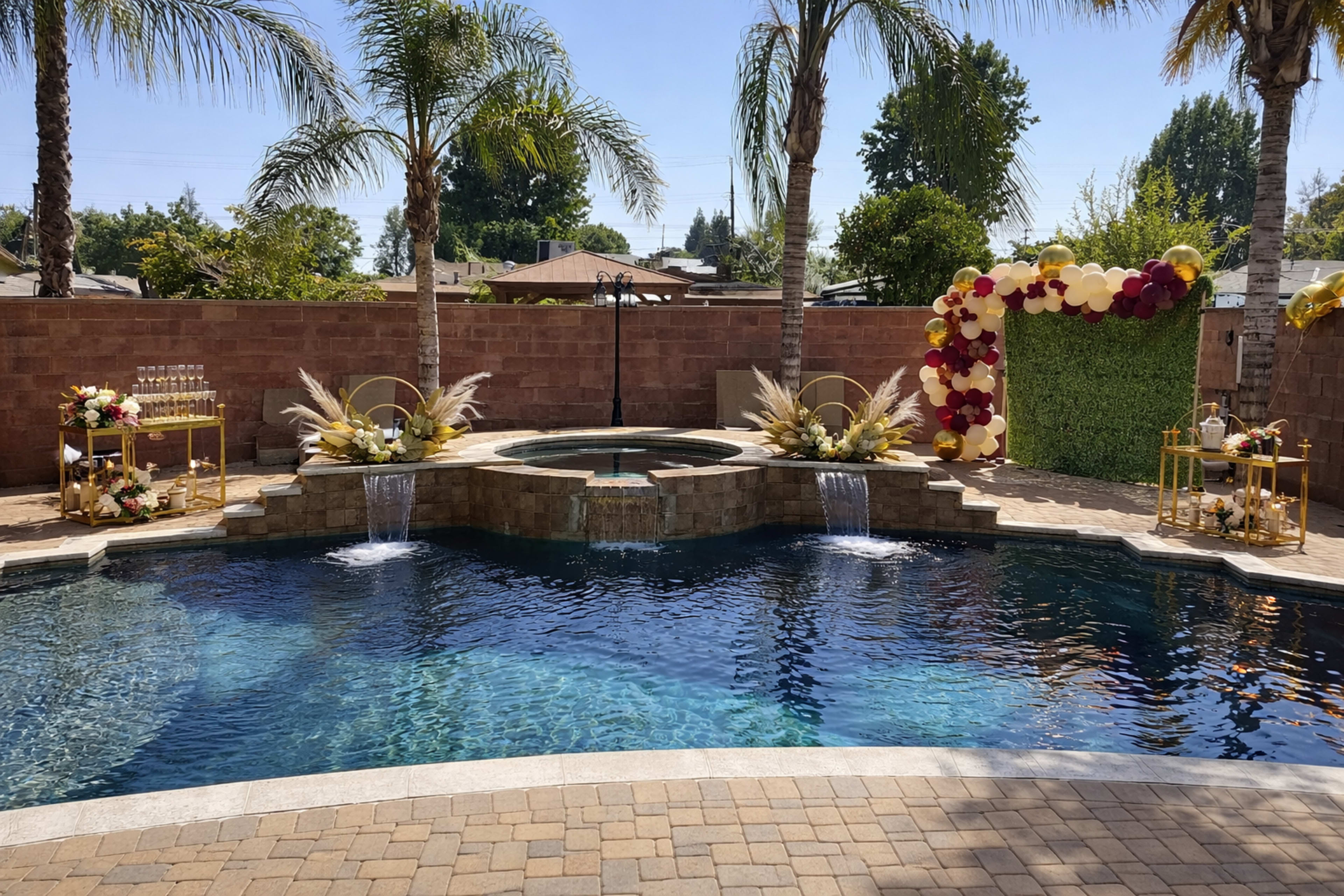 A luxurious backyard features a swimming pool with waterfalls, surrounded by palm trees, and is adorned with decorative elements for an event that includes floral arrangements and a vibrant arch.