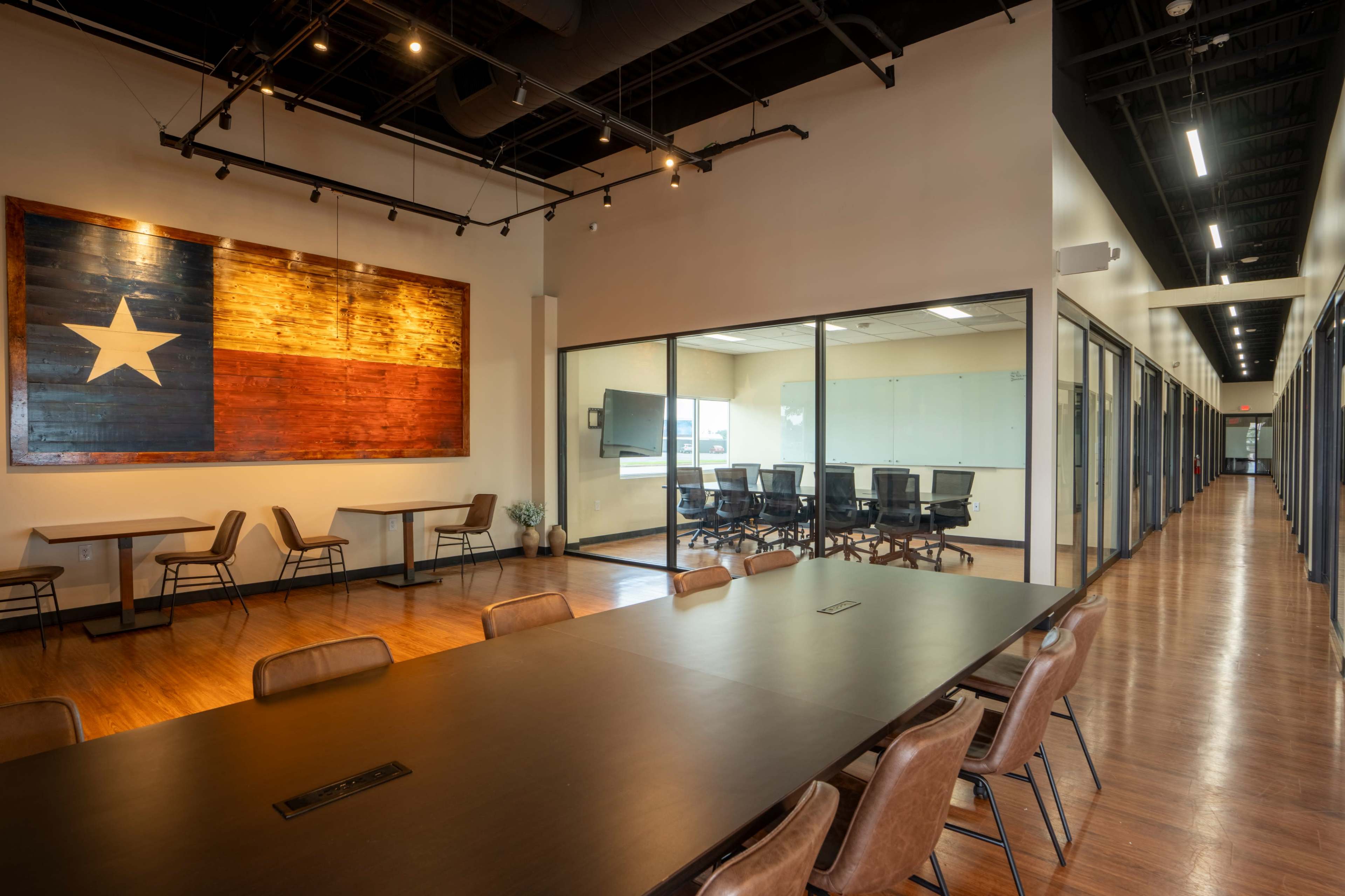 A spacious modern office features a long table and chairs, with a Texas flag mural on the wall and glass-walled meeting rooms along a hallway.