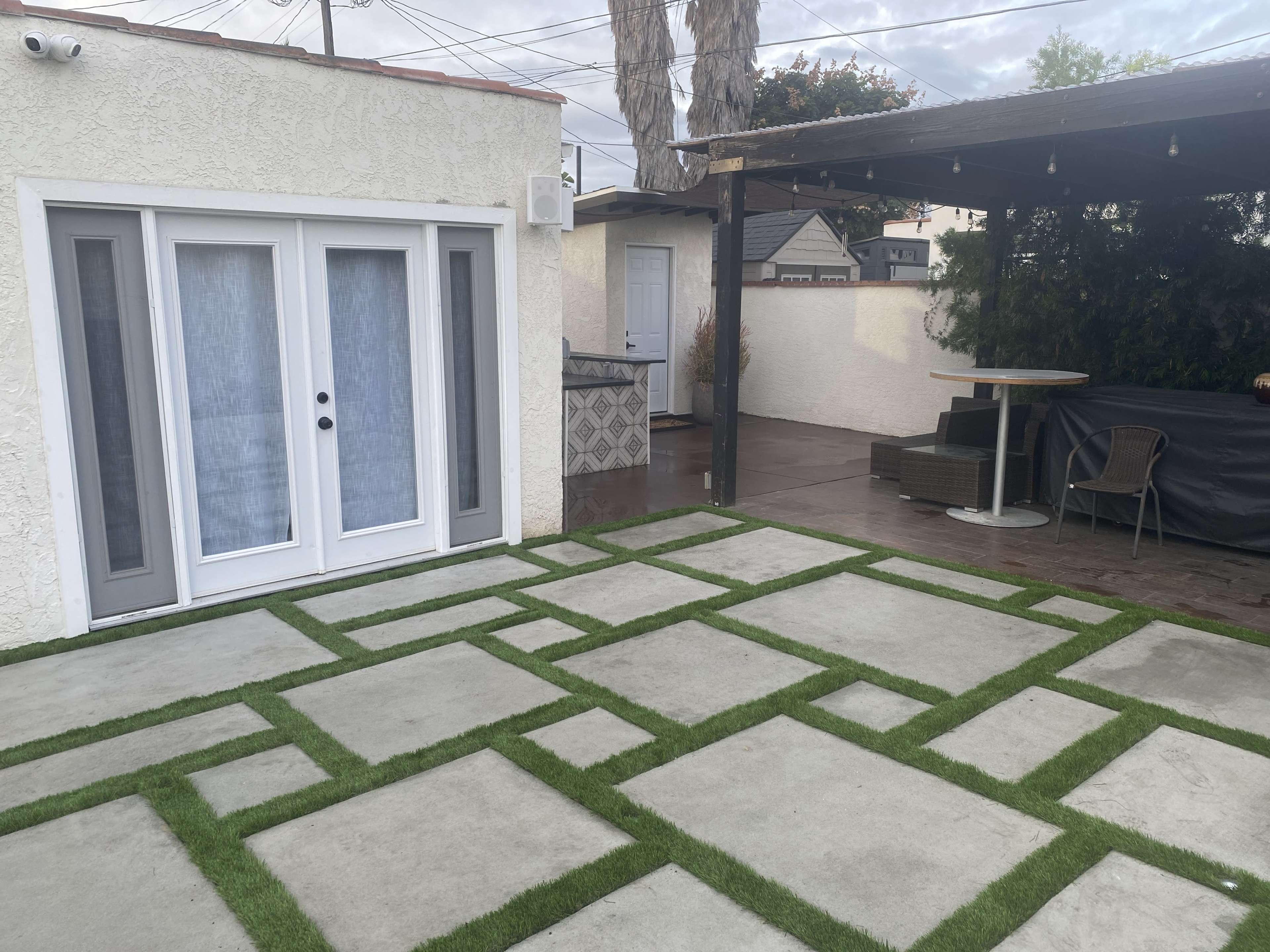 A patio area featuring a concrete and grass pattern, with an outdoor seating space and a small building in the background.