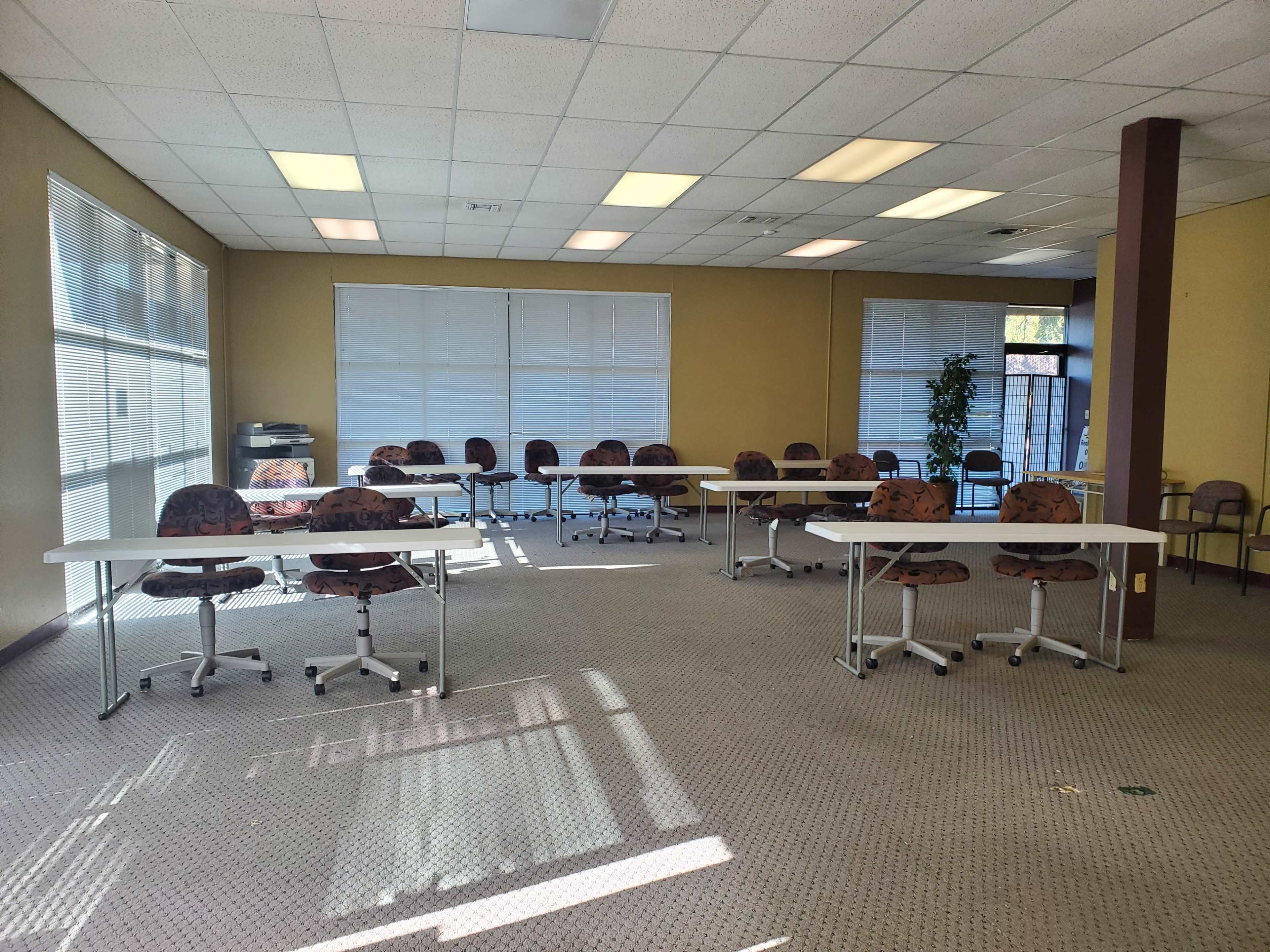 A spacious room with several rows of tables and chairs arranged for a meeting or training session, illuminated by natural light through large windows.