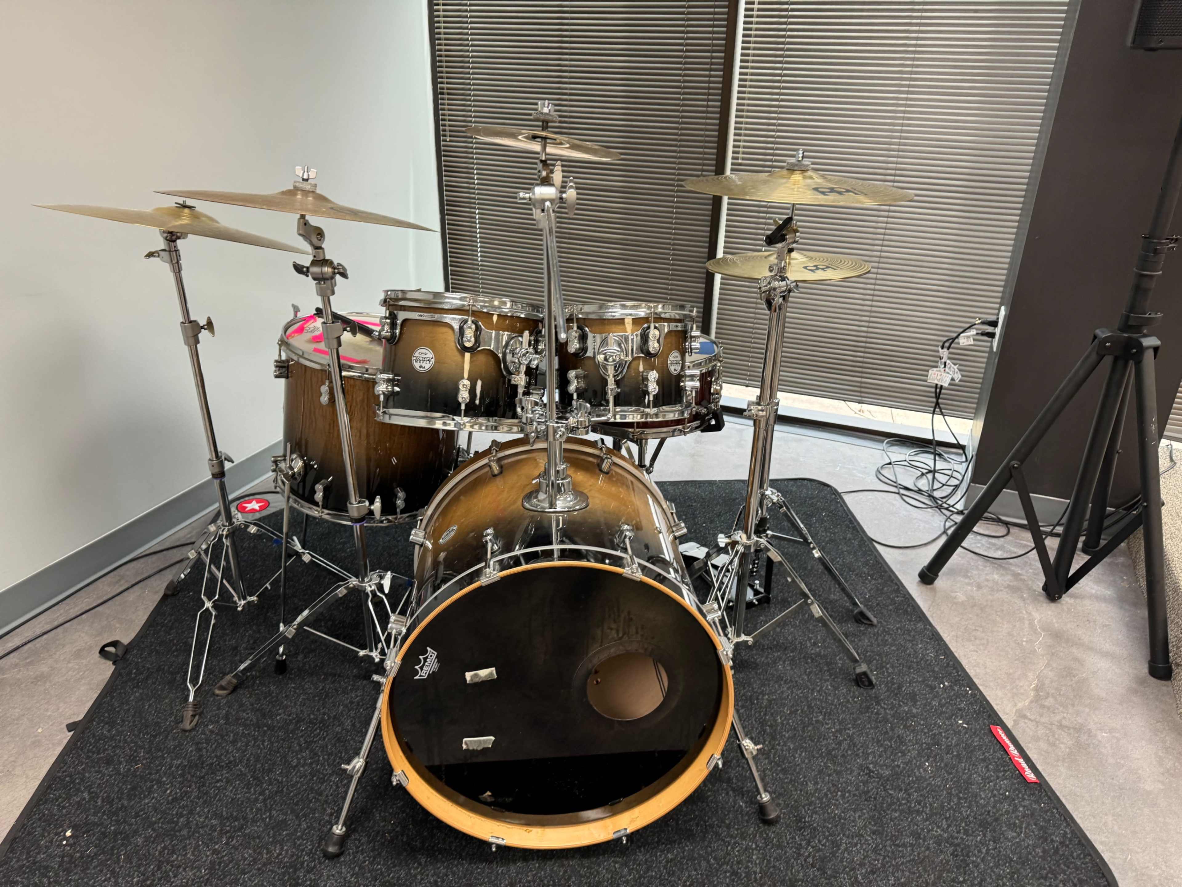 The image shows a drum set positioned on a black mat in a room with gray walls and large windows.