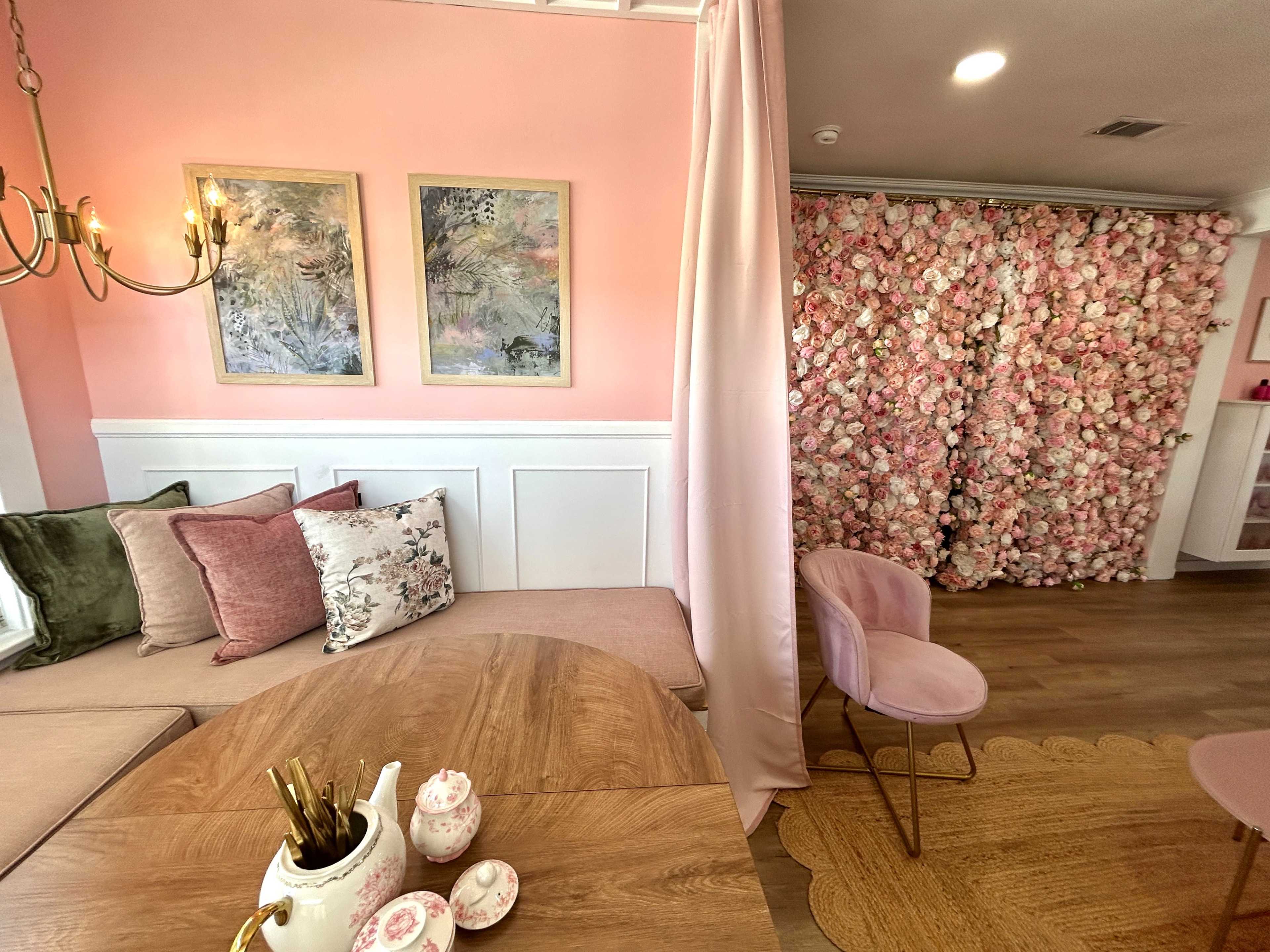The image shows a cozy interior with pink walls, floral artwork, a wooden table, plush seating, and a decorative flower wall at the back.