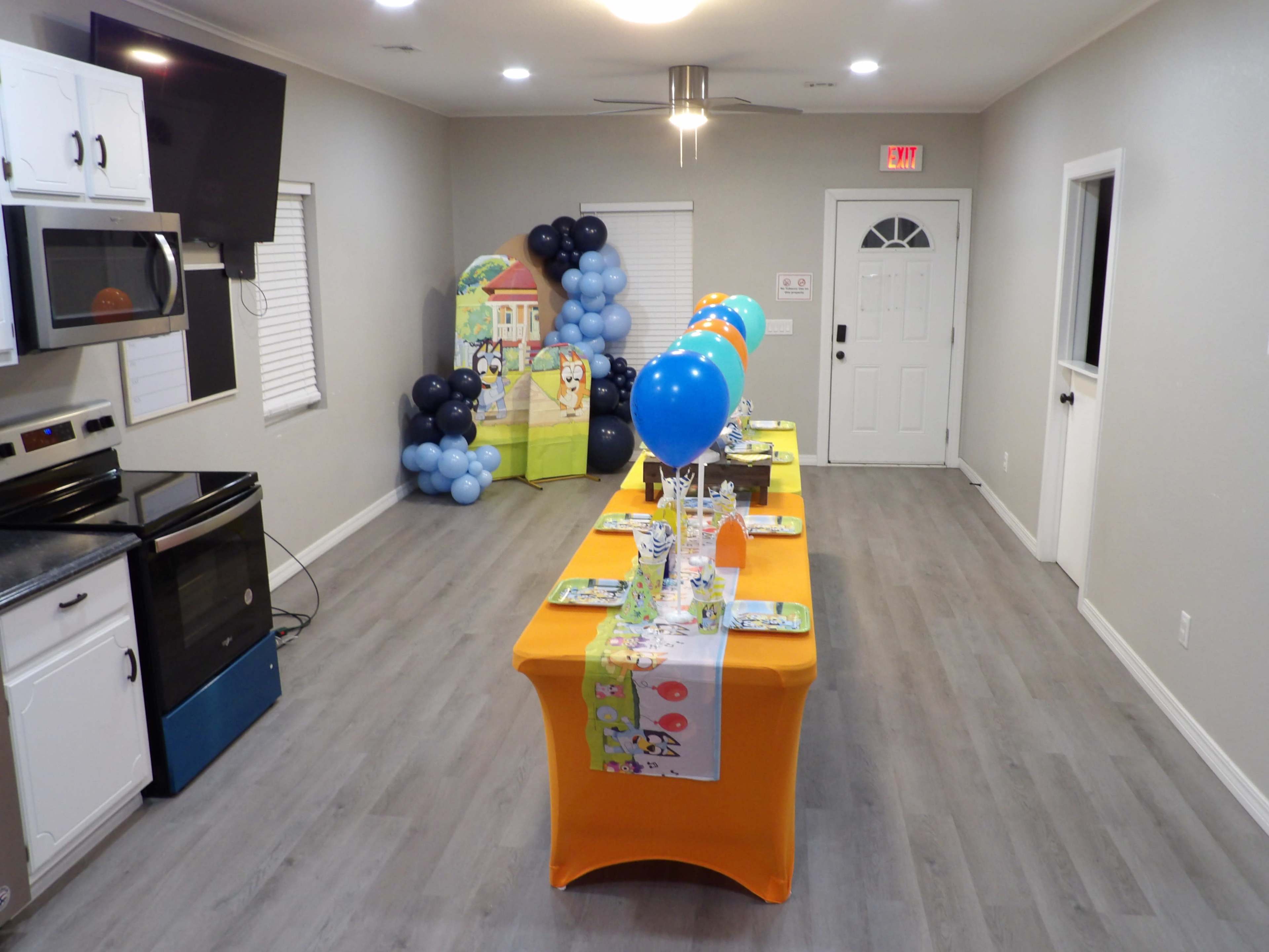 A party setup is displayed in a room featuring a long table covered with an orange tablecloth, decorated with colorful balloons and party supplies, against a backdrop of a kitchen and an entrance door.