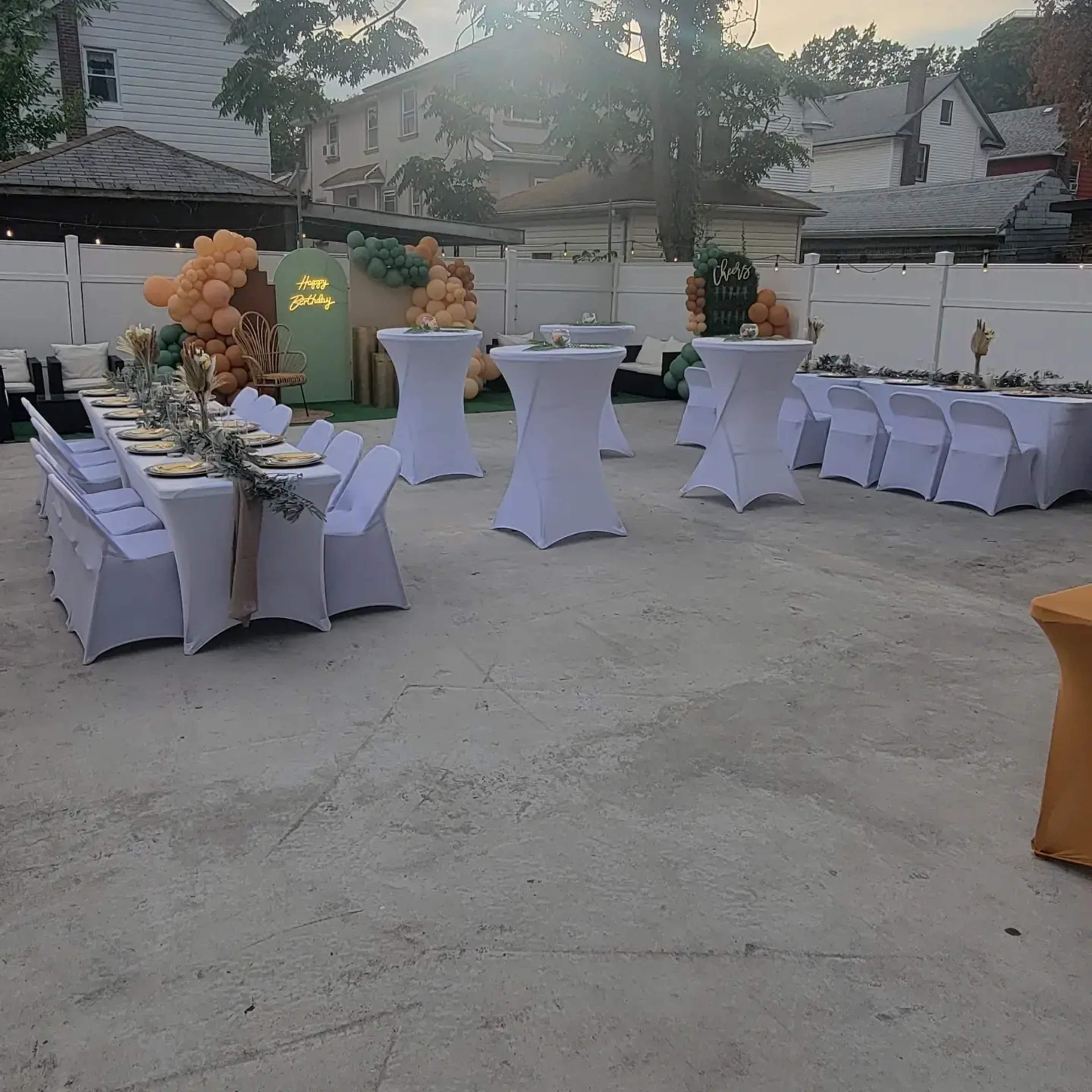 The image shows an outdoor event setup with several tables covered in white linens, decorated with balloons and greenery in a patio area.