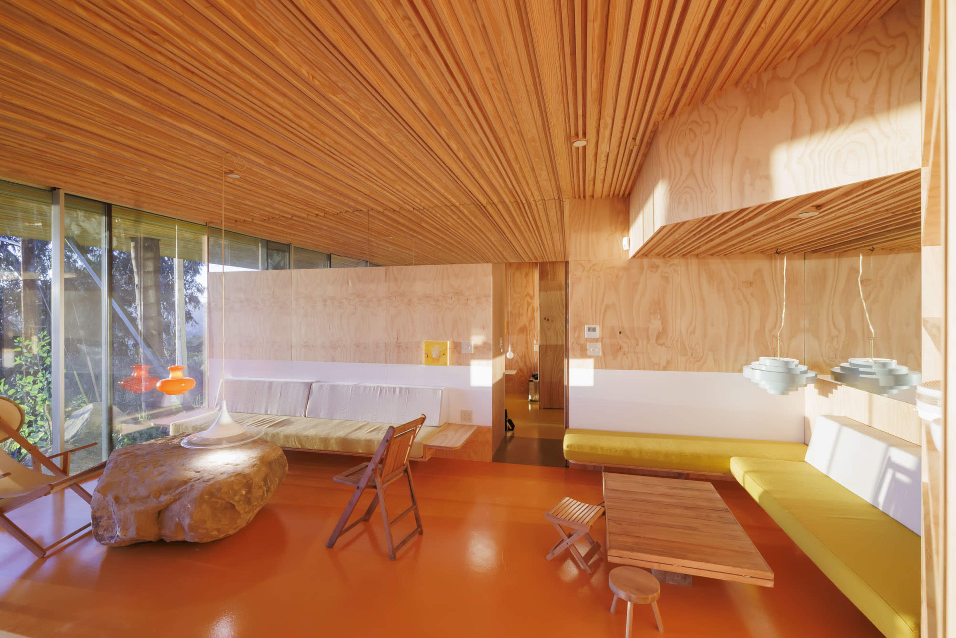 The image shows a modern, minimalist interior with wooden paneling, bright orange flooring, and a mix of furniture including a stone table, a yellow bench, and various light fixtures.
