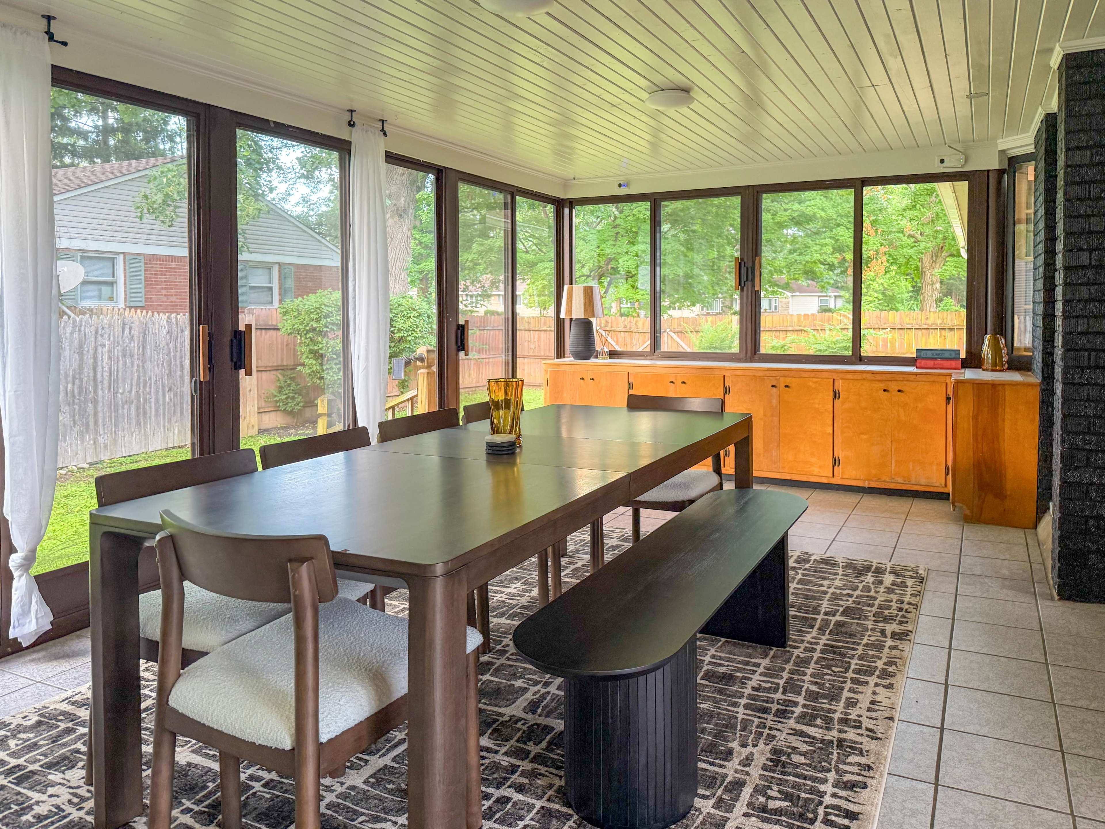 The image shows a spacious sunroom with a large dining table and benches, large windows, and wooden cabinetry against a backdrop of greenery outside.
