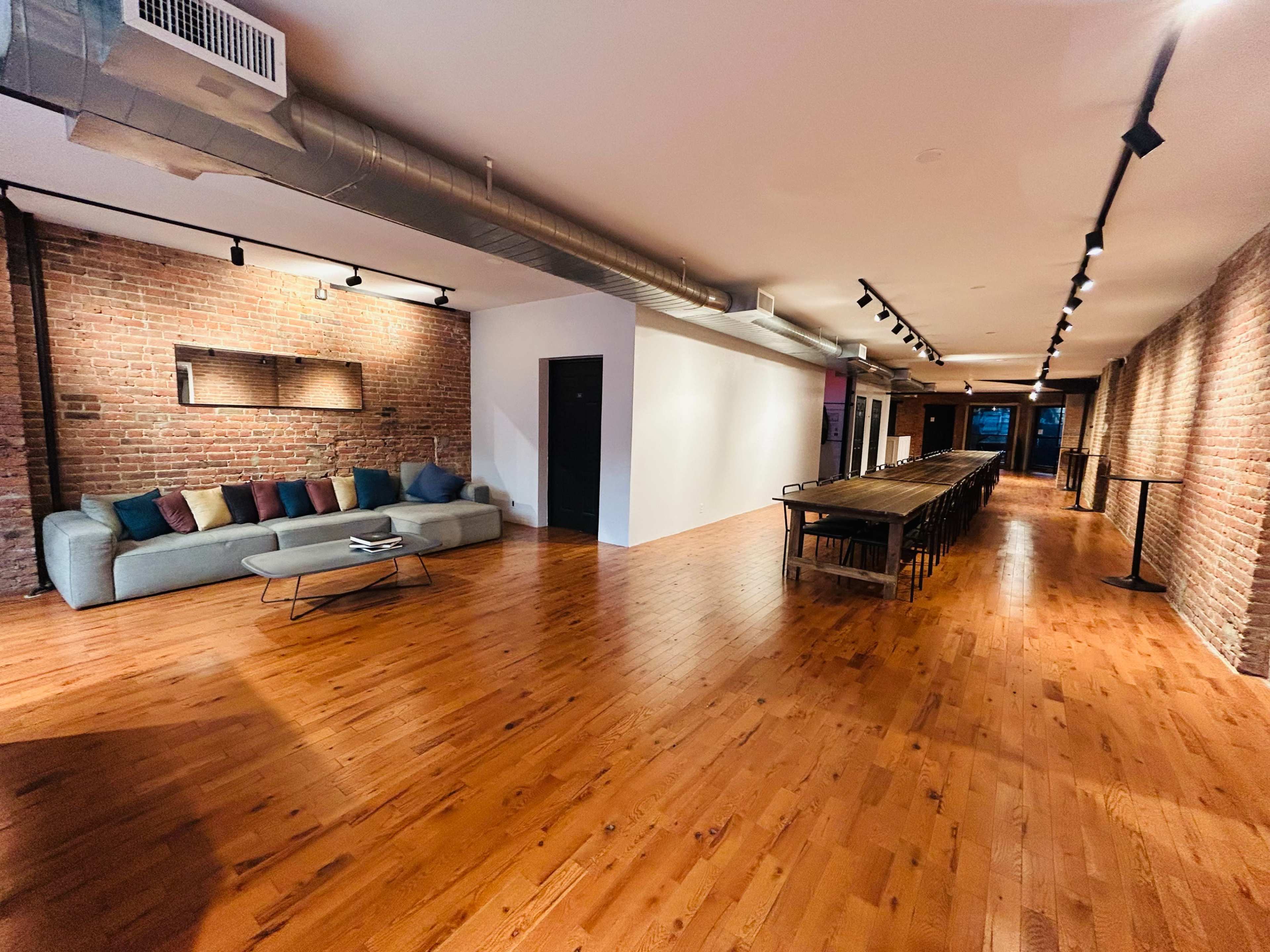 The image shows a spacious room with wooden flooring, exposed brick walls, a long dining table, and a sofa with colorful cushions.