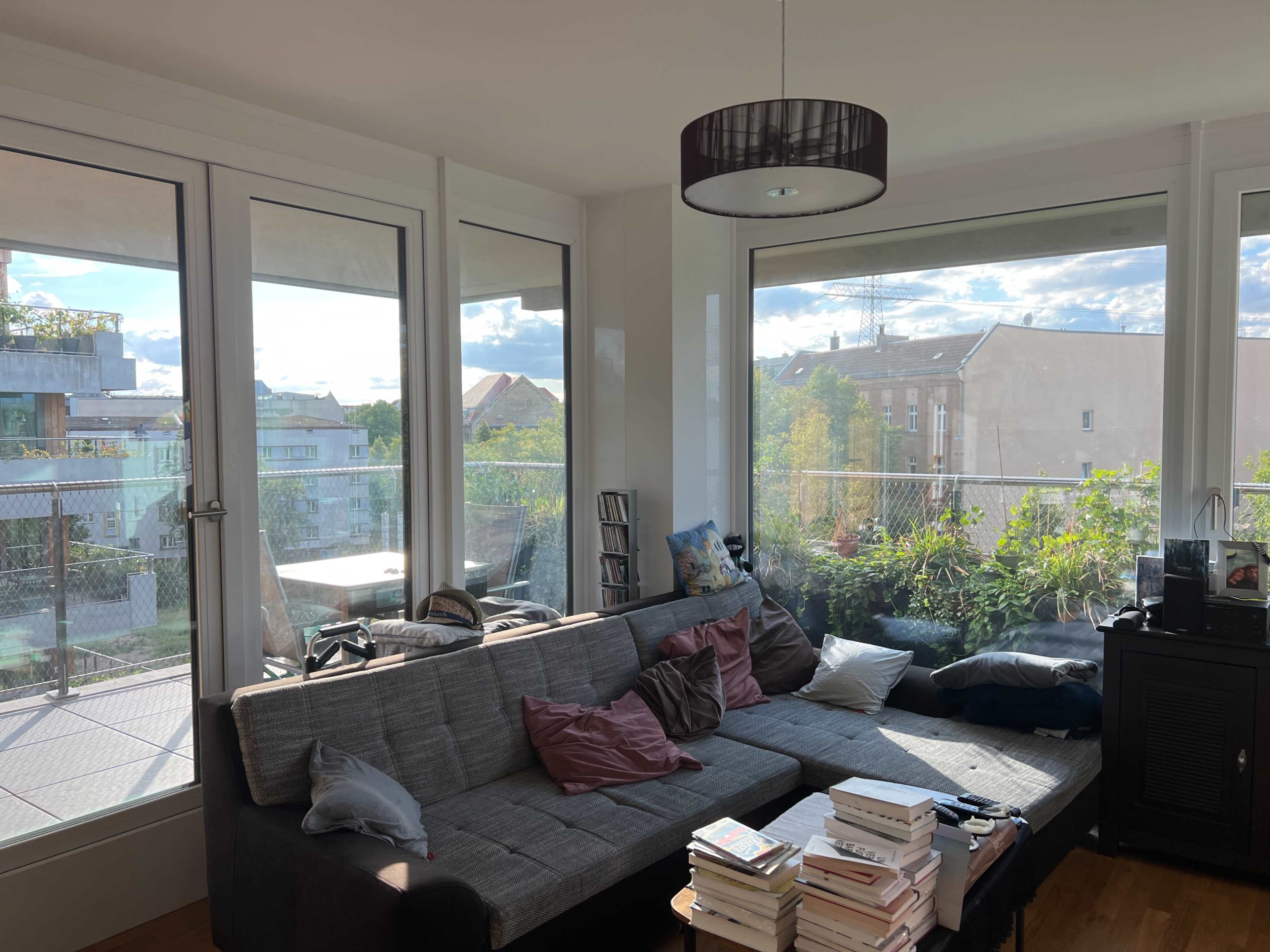 A cozy living room with large windows that overlook a balcony and greenery outside, featuring a gray sofa adorned with pillows and a coffee table stacked with books.