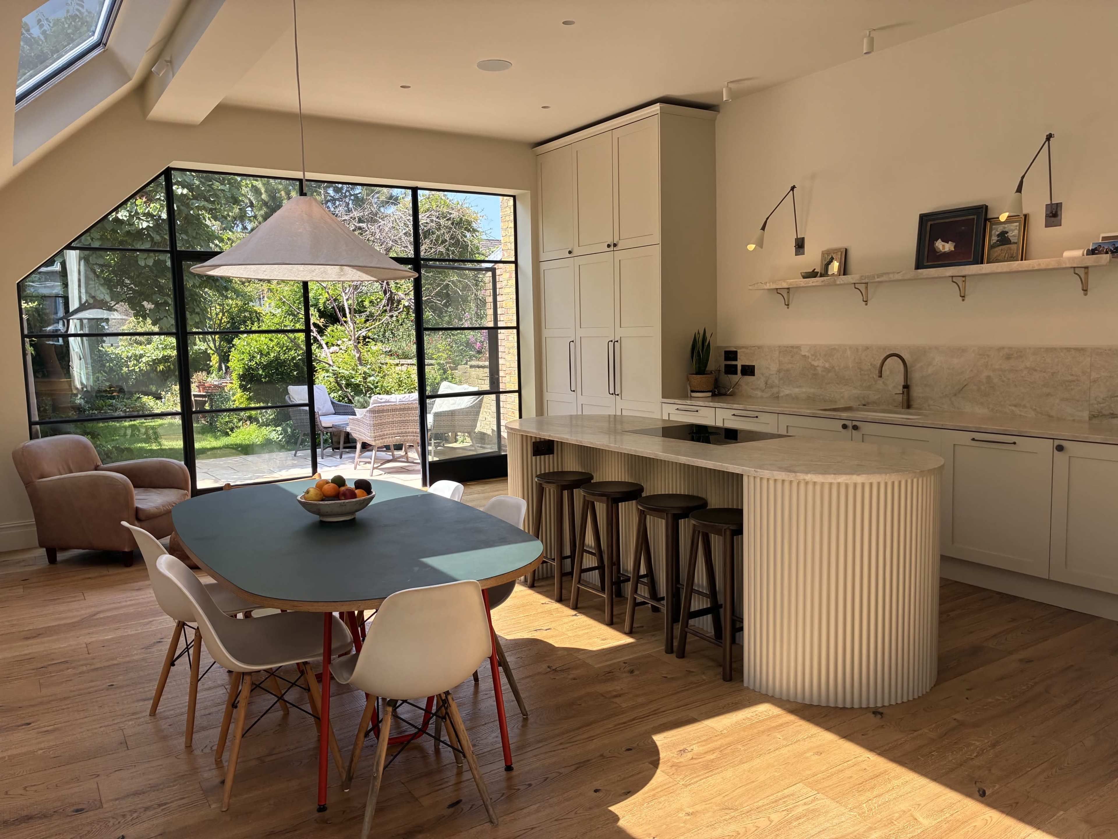 The image shows a modern kitchen with a round dining table, bar stools, and large windows overlooking a garden.