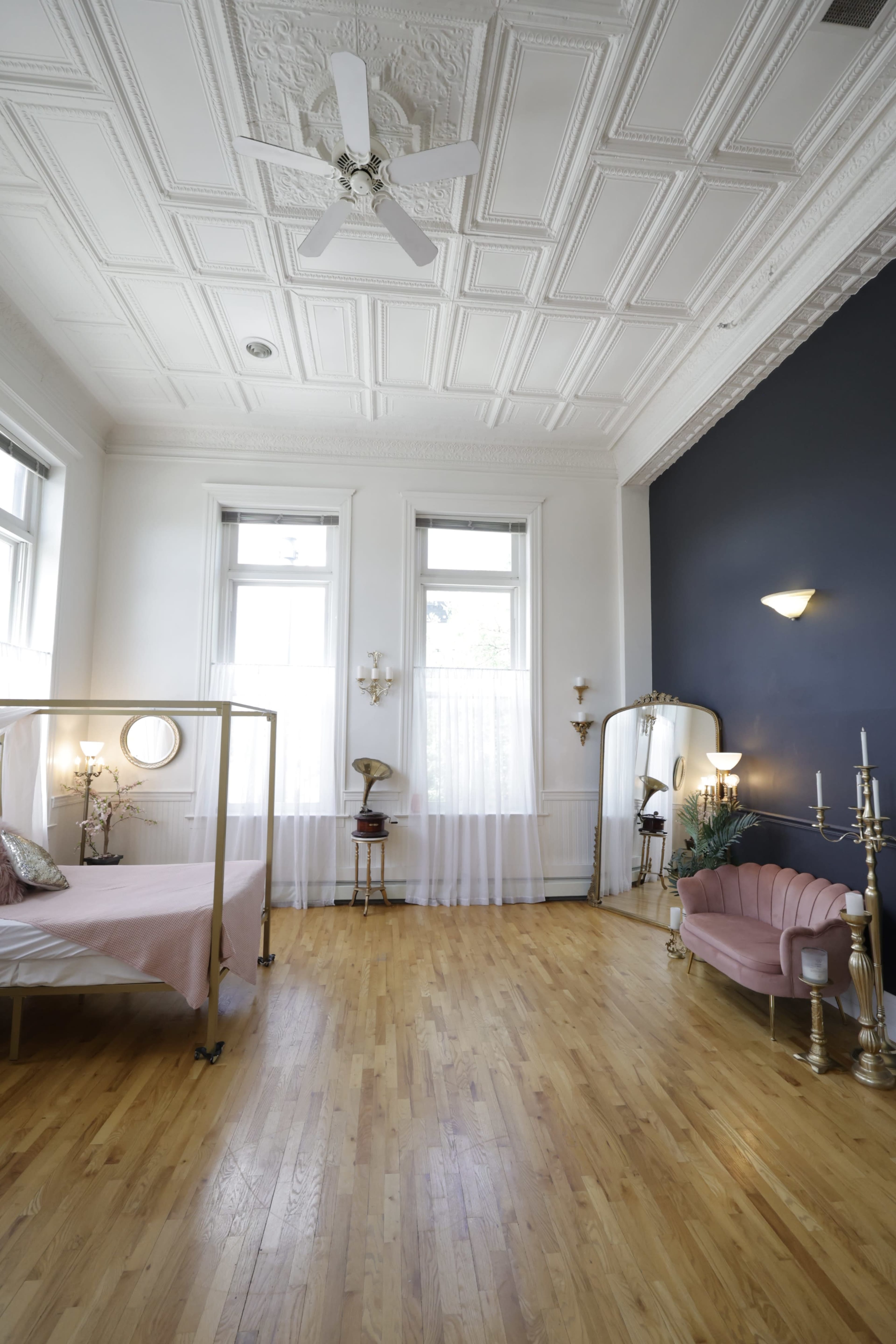 The image shows a spacious, well-lit bedroom featuring a bed with a pink bedspread, a vintage mirror, and elegant wall sconces against a dark accent wall.
