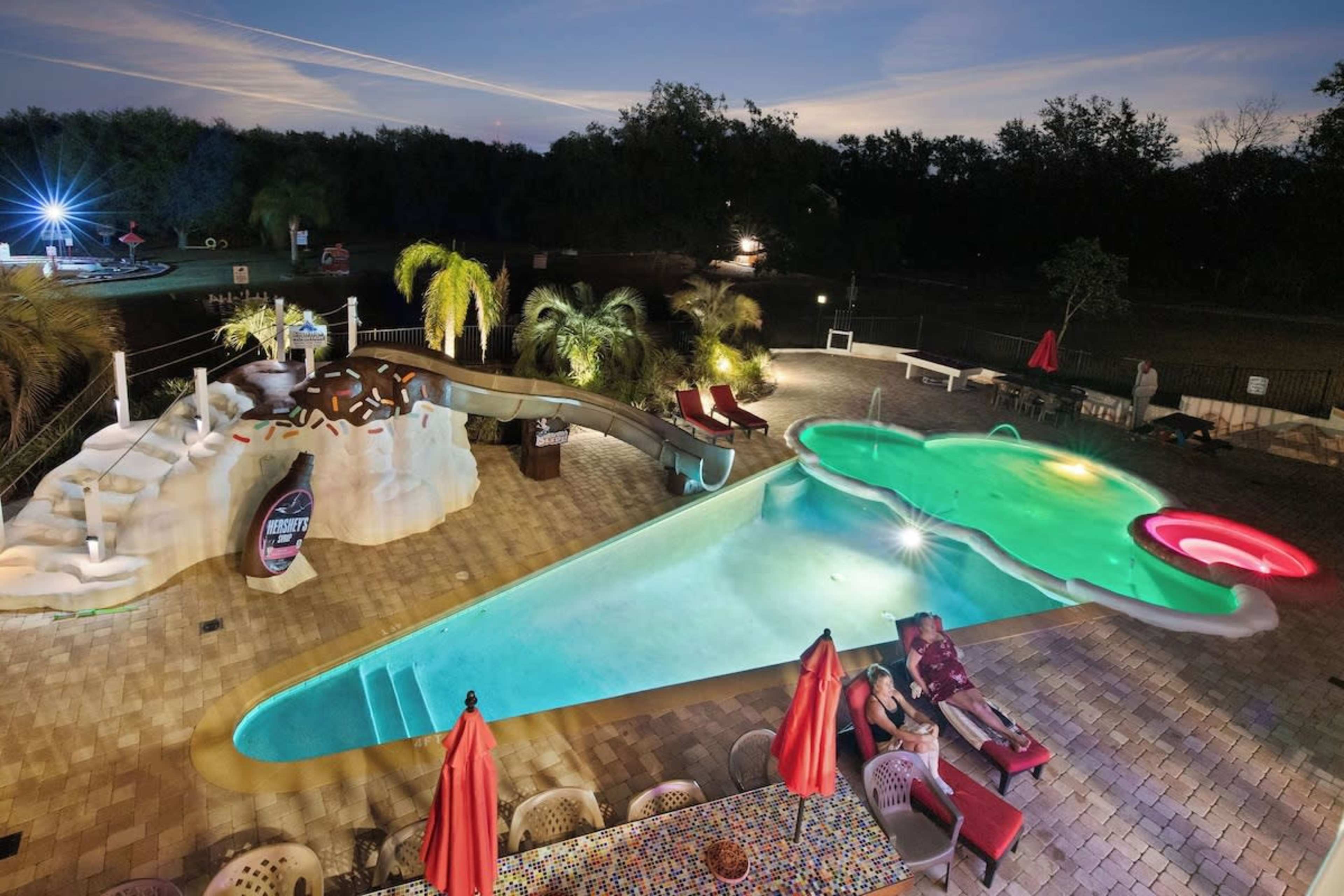 The scene features a large, uniquely shaped swimming pool with colorful slides and surrounded by palm trees and lounge chairs under a twilight sky.