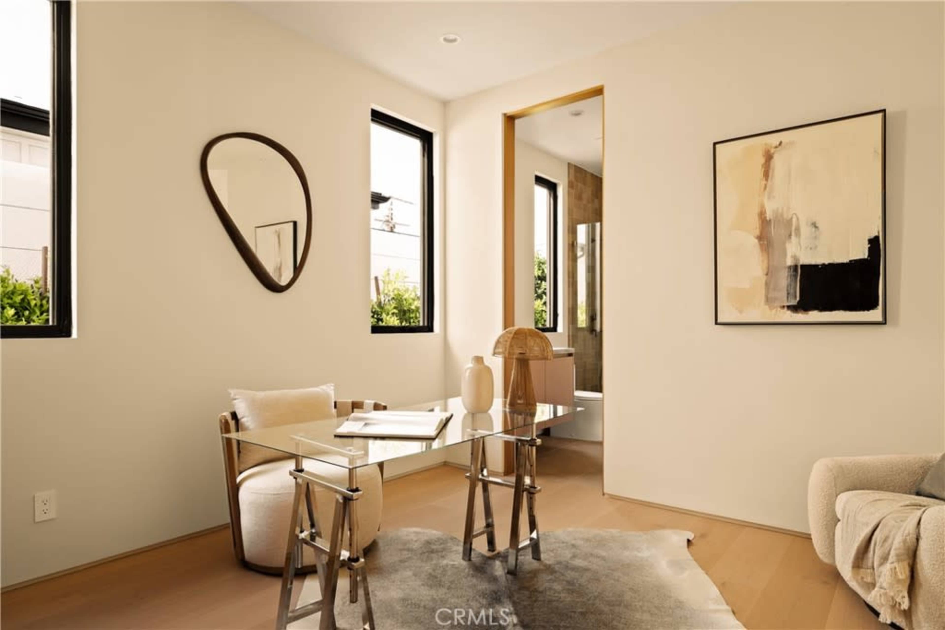 The image shows a modern interior space featuring a small glass table with a chair, a round mirror on the wall, and a large artwork, all surrounded by large windows.