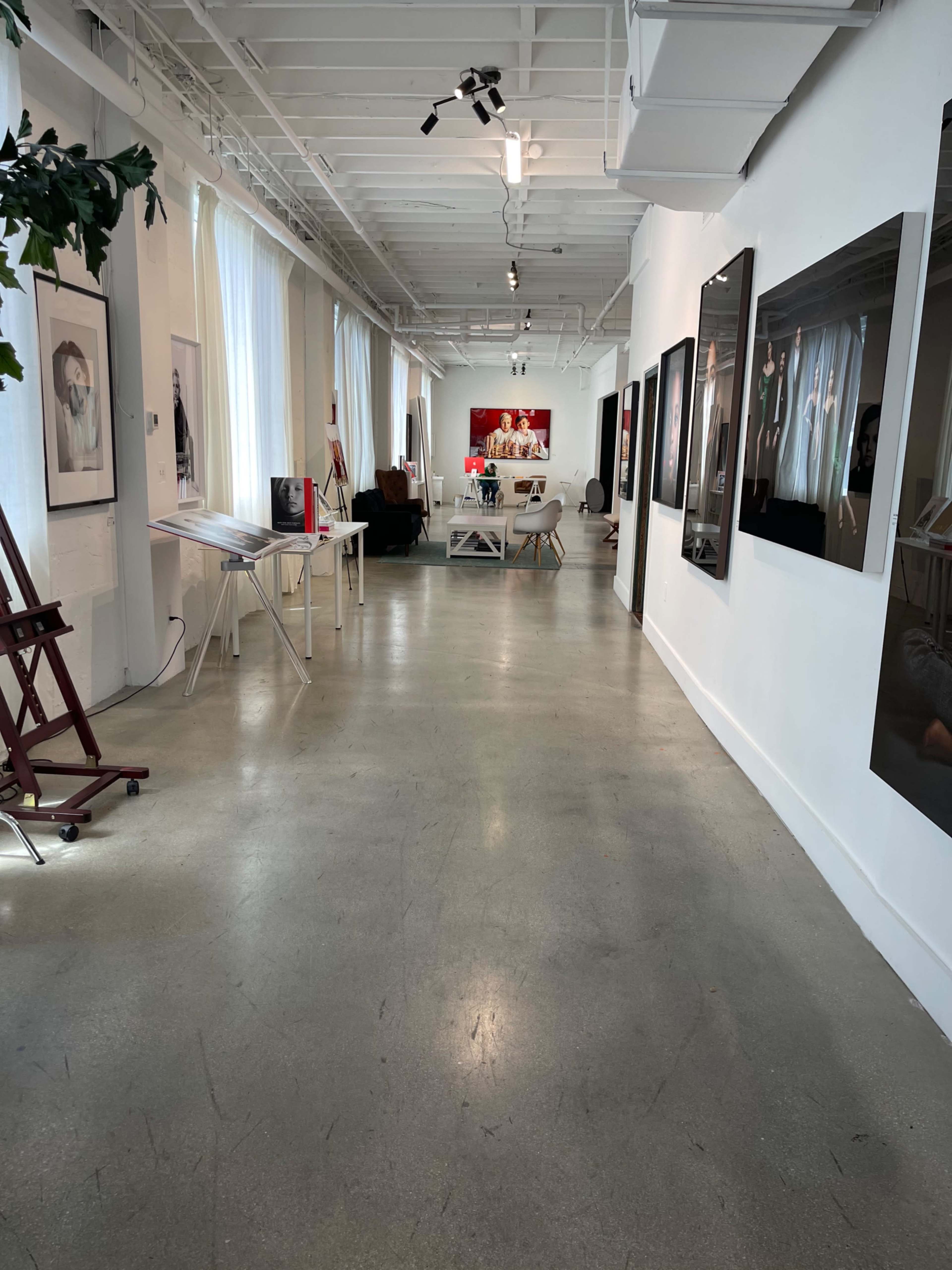 The image shows an art gallery with a long, narrow hallway featuring various artworks on the walls and a seating area at the end.