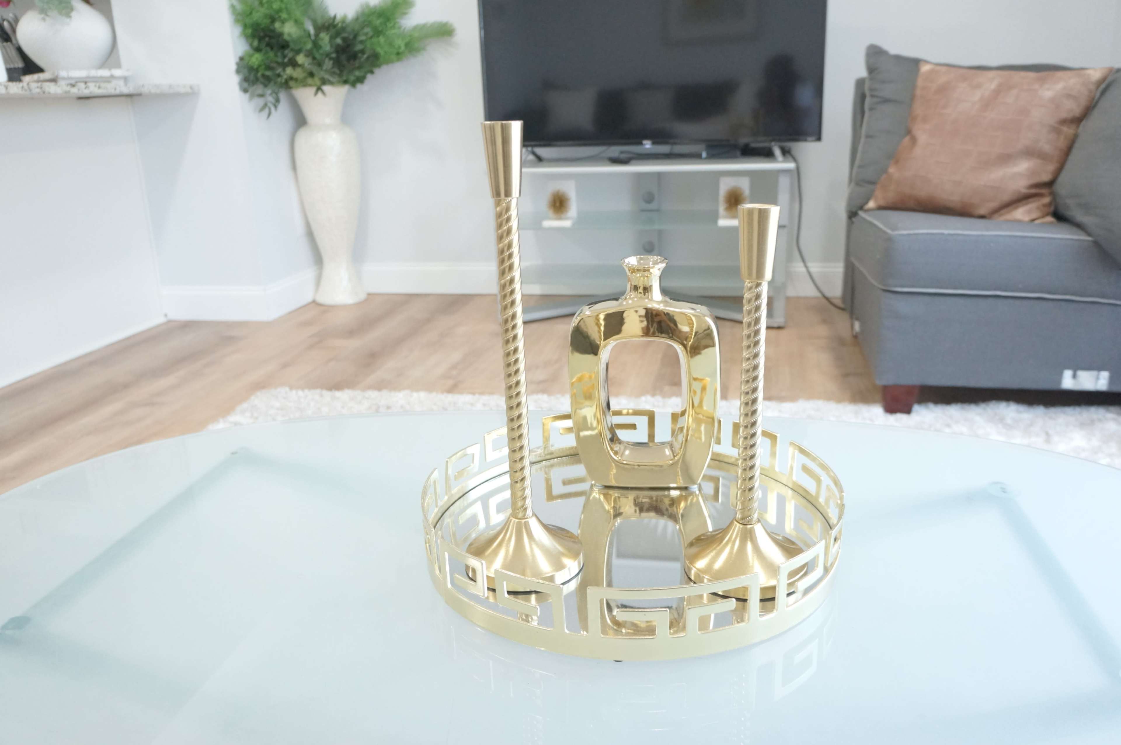 A golden decorative tray with two tall candlesticks and a small decorative bottle is displayed on a glass coffee table in a living room.