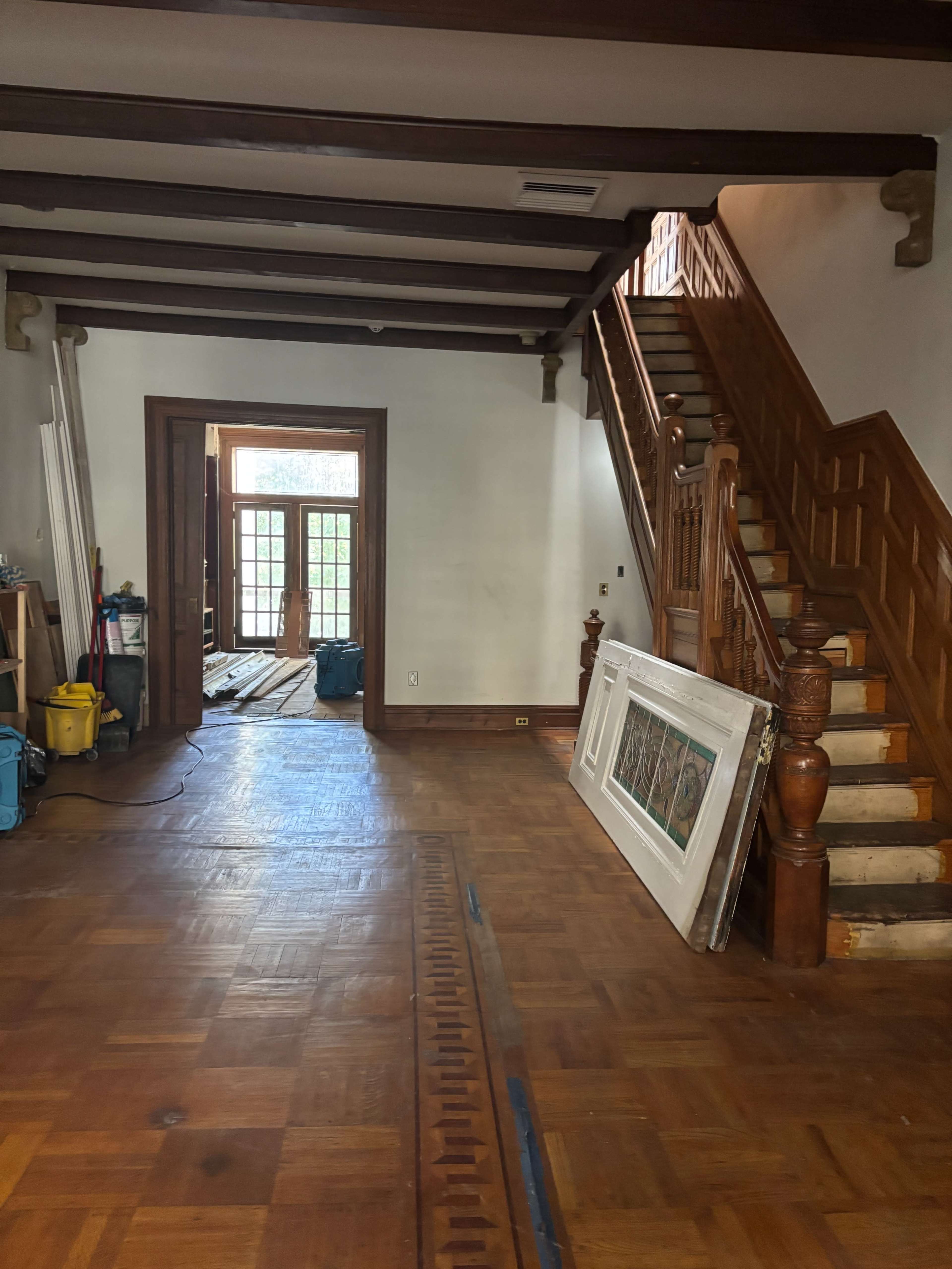 The image shows the interior of a partially furnished room with wooden flooring, a staircase leading to an upper level, and a doorway leading to another room.