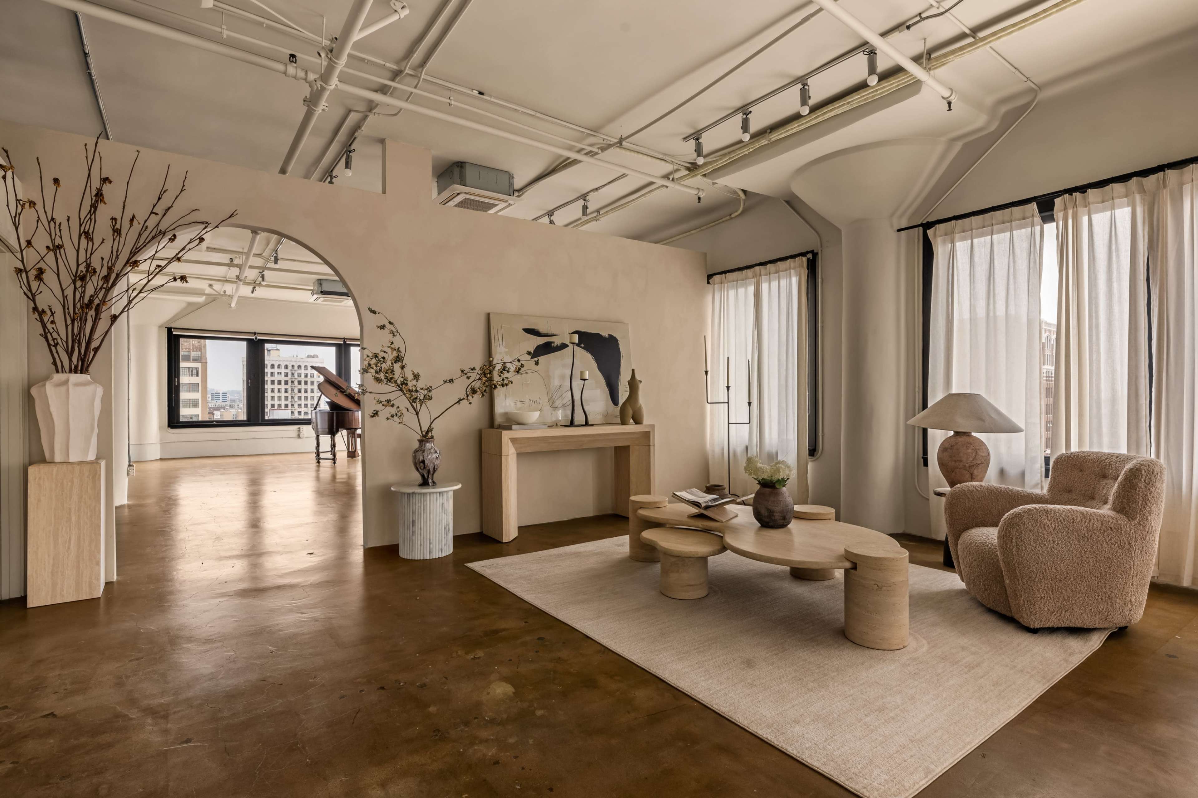 The image shows a modern interior design featuring a lounge area with a circular wooden coffee table, a plush chair, and large windows framed by sheer curtains.