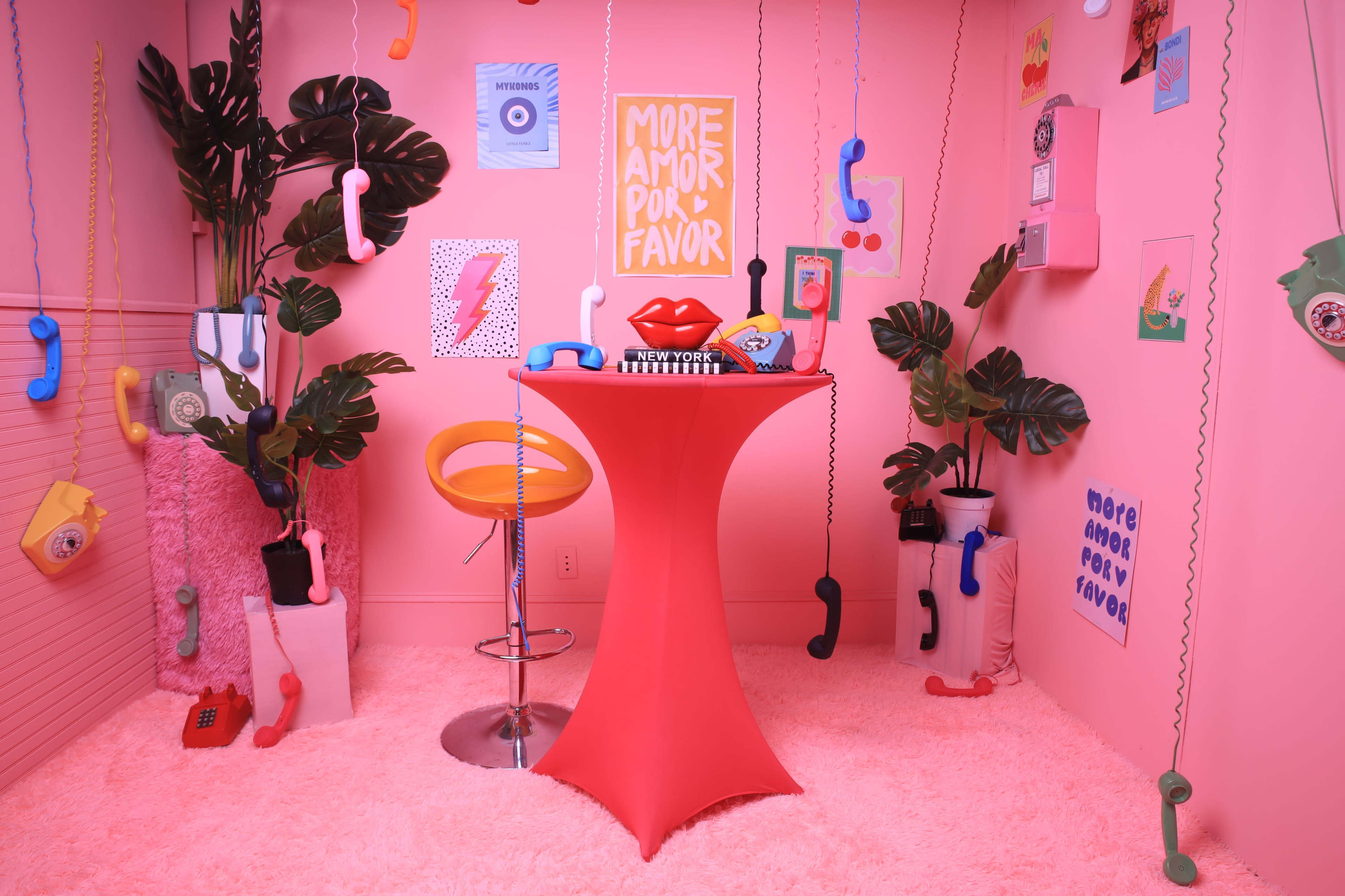 The image features a vibrant pink room decorated with various decorations, including colorful telephones hanging from the ceiling, a sleek orange bar stool, and a red table with lip-shaped decor.