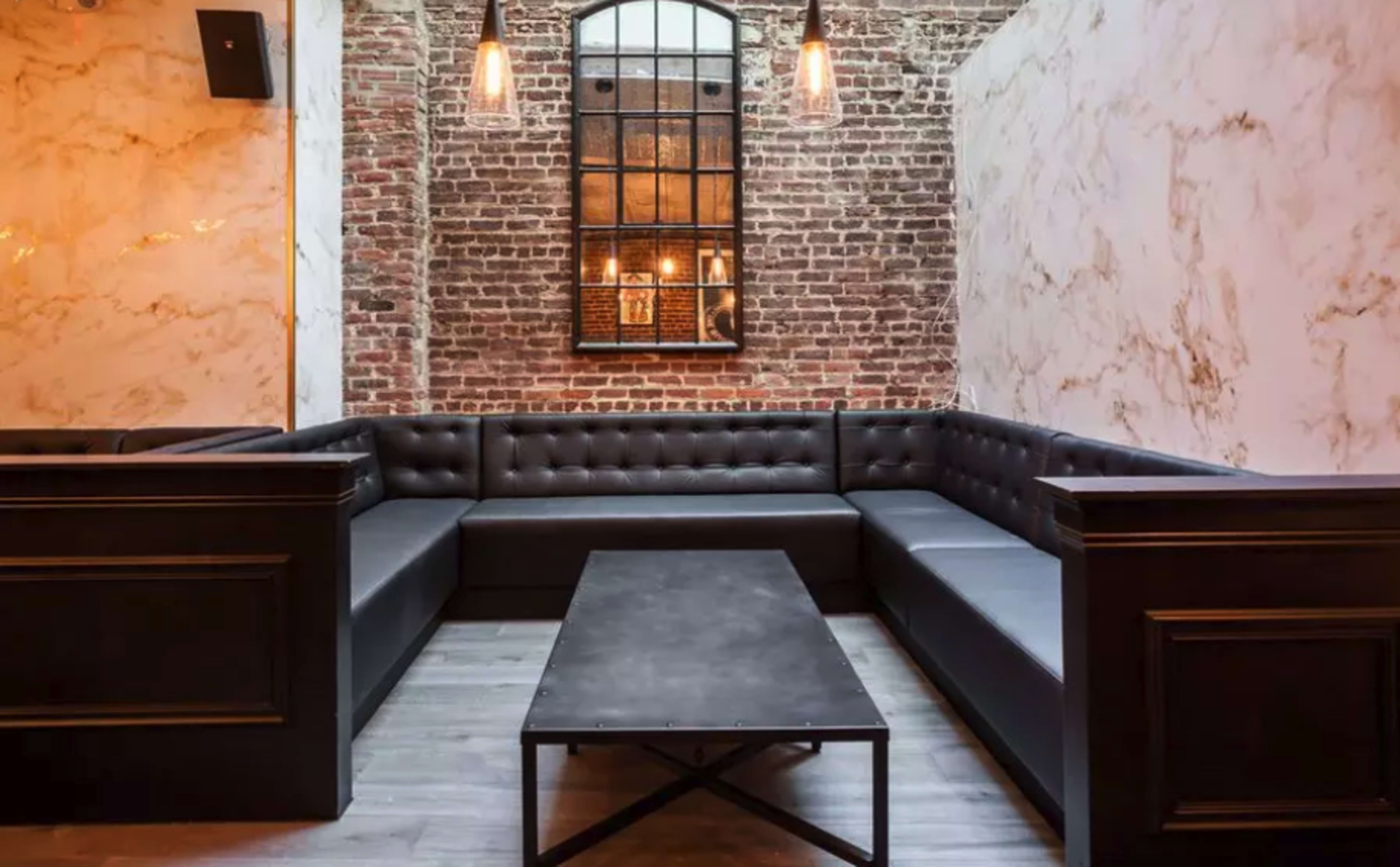 The image depicts a modern, minimalist lounge area featuring a large, black sectional sofa and a rectangular coffee table set against a backdrop of exposed brick walls and elegant marble accents.