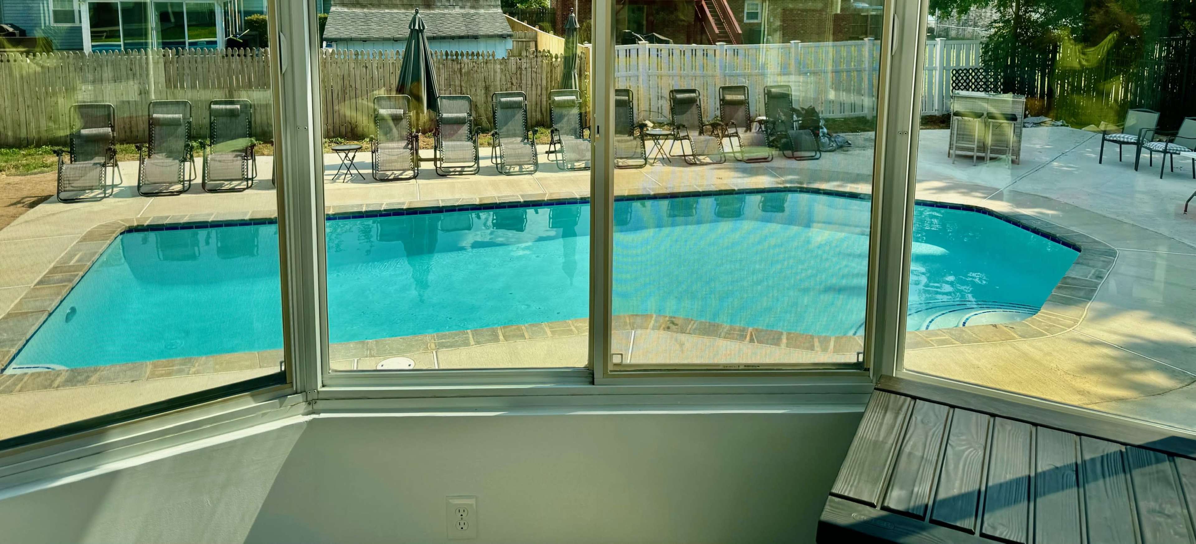A view through a large window of a swimming pool surrounded by loungers and a patio area.