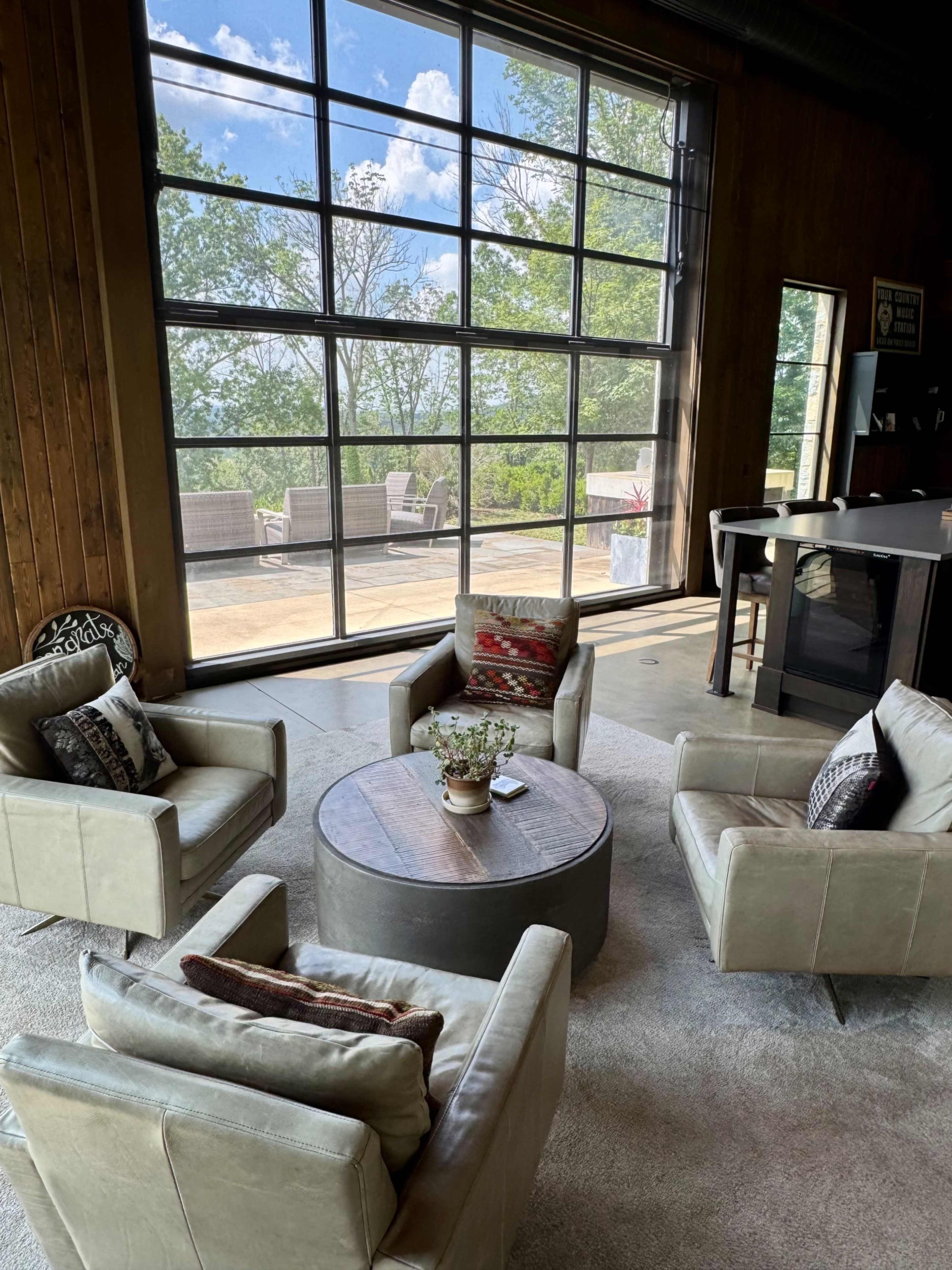 The image shows a modern living space with four armchairs arranged around a circular wooden table, facing large windows that overlook a green landscape.