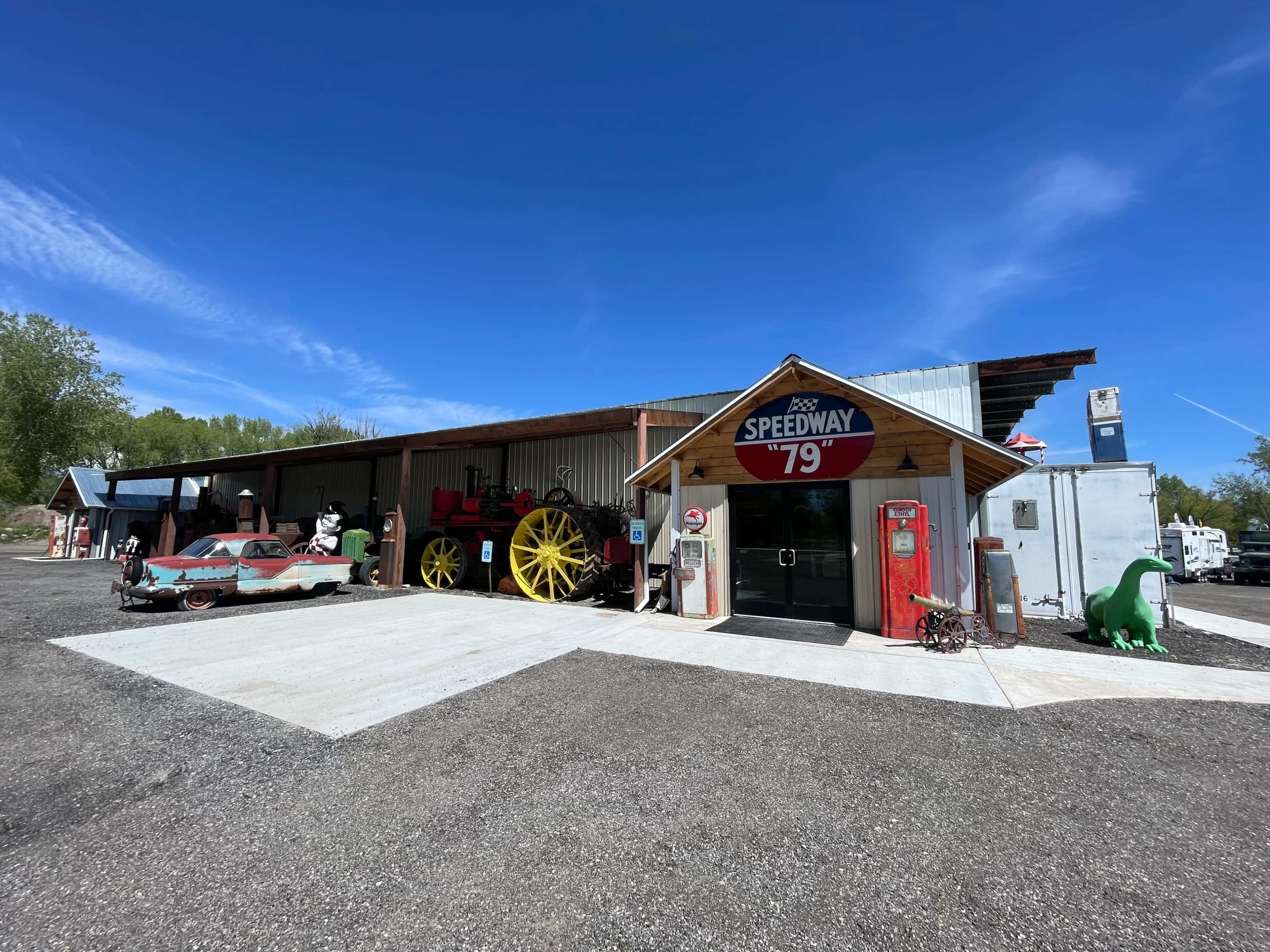 A vintage storefront marked "Speedway 79" displays classic cars and memorabilia alongside a large yellow wagon in a rural setting.
