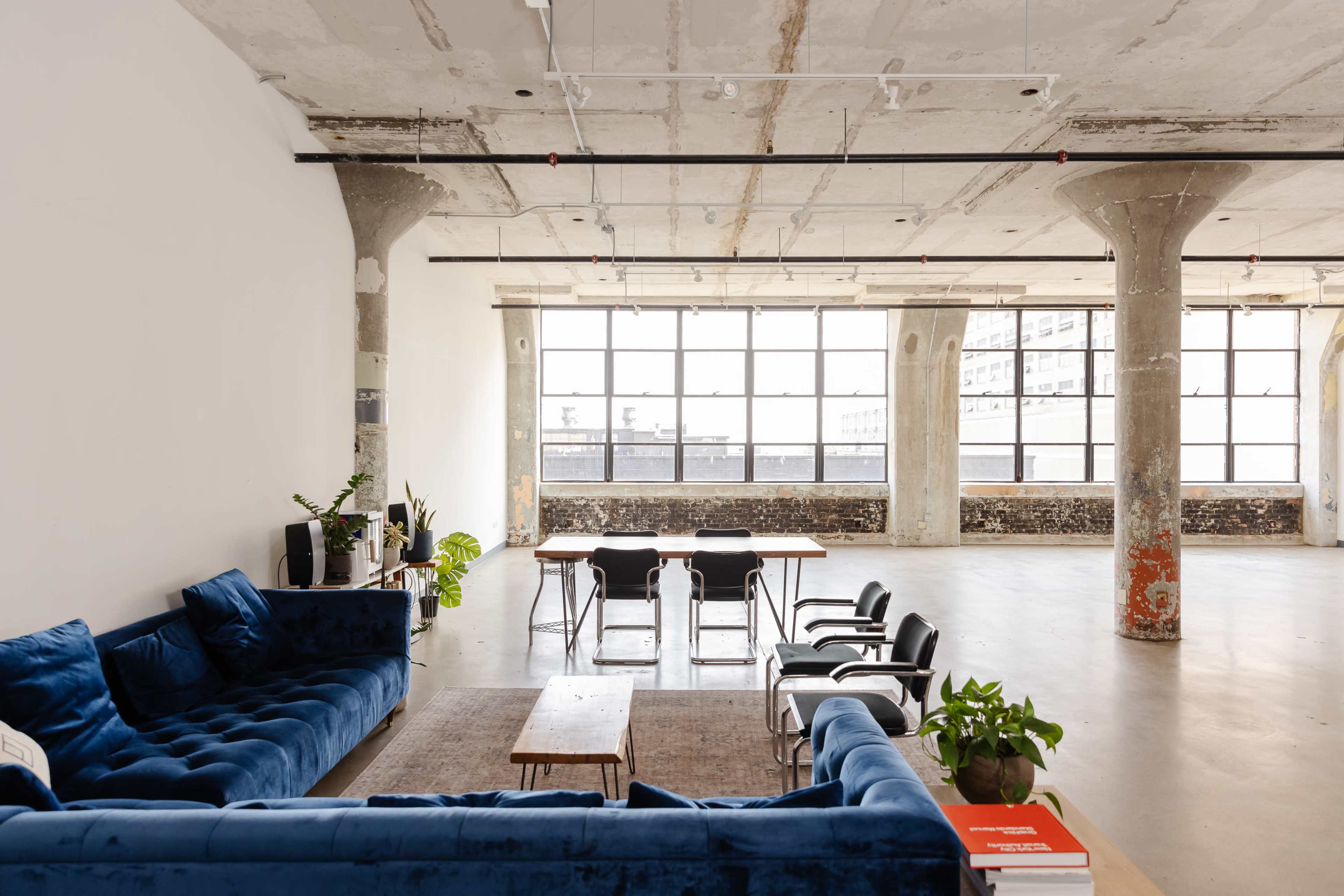 A spacious, industrial-style room features a blue sofa, a wooden coffee table, a dining table with chairs, and large windows that illuminate the space.